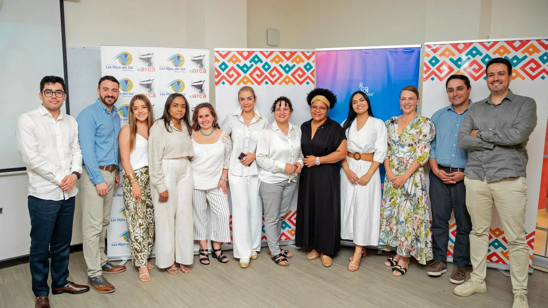 Grupo de personas posando delante banners institucionales de los proyectos "Los Hijos del Sol" y "el arca". Todos visten de manera formal y casual, formando una fila en un espacio cerrado, posiblemente en un evento académico o de presentación.