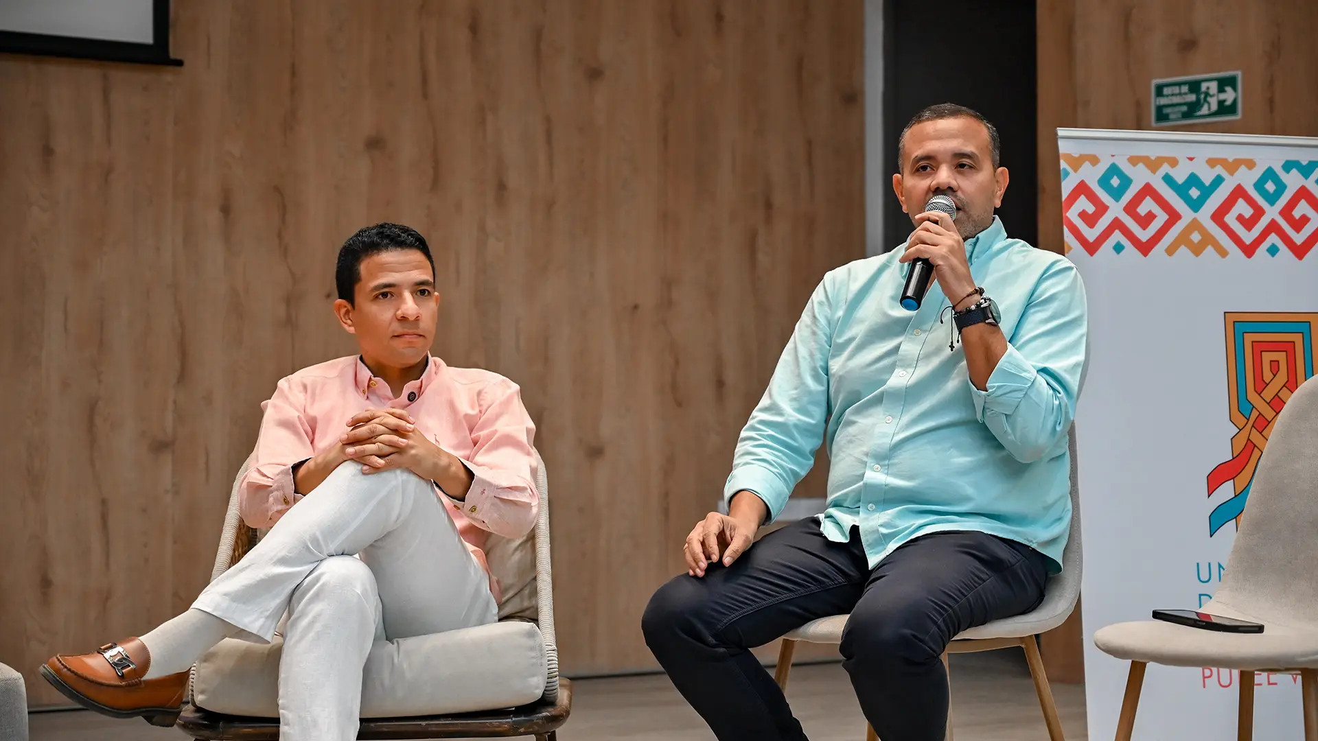Dos panelistas participan en un conversatorio académico; uno de ellos toma la palabra con micrófono en mano, mientras el otro escucha con atención. Ambos están sentados en un auditorio con fondo de madera y elementos gráficos institucionales.