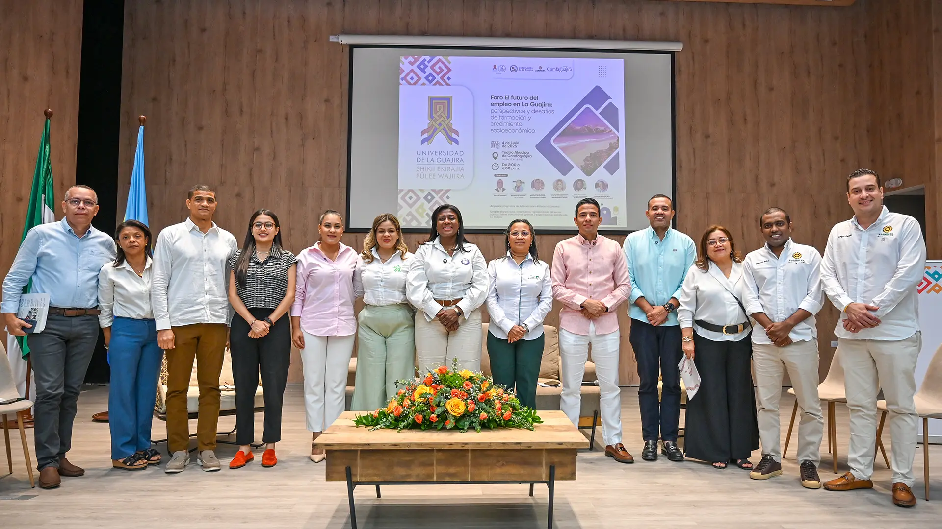 Grupo de trece personas posando para una foto grupal en un auditorio. Están frente a una pantalla que muestra el título del foro: “El futuro del empleo en La Guajira”. El evento se desarrolla en la Universidad de La Guajira. Las personas están vestidas de manera formal y elegante.