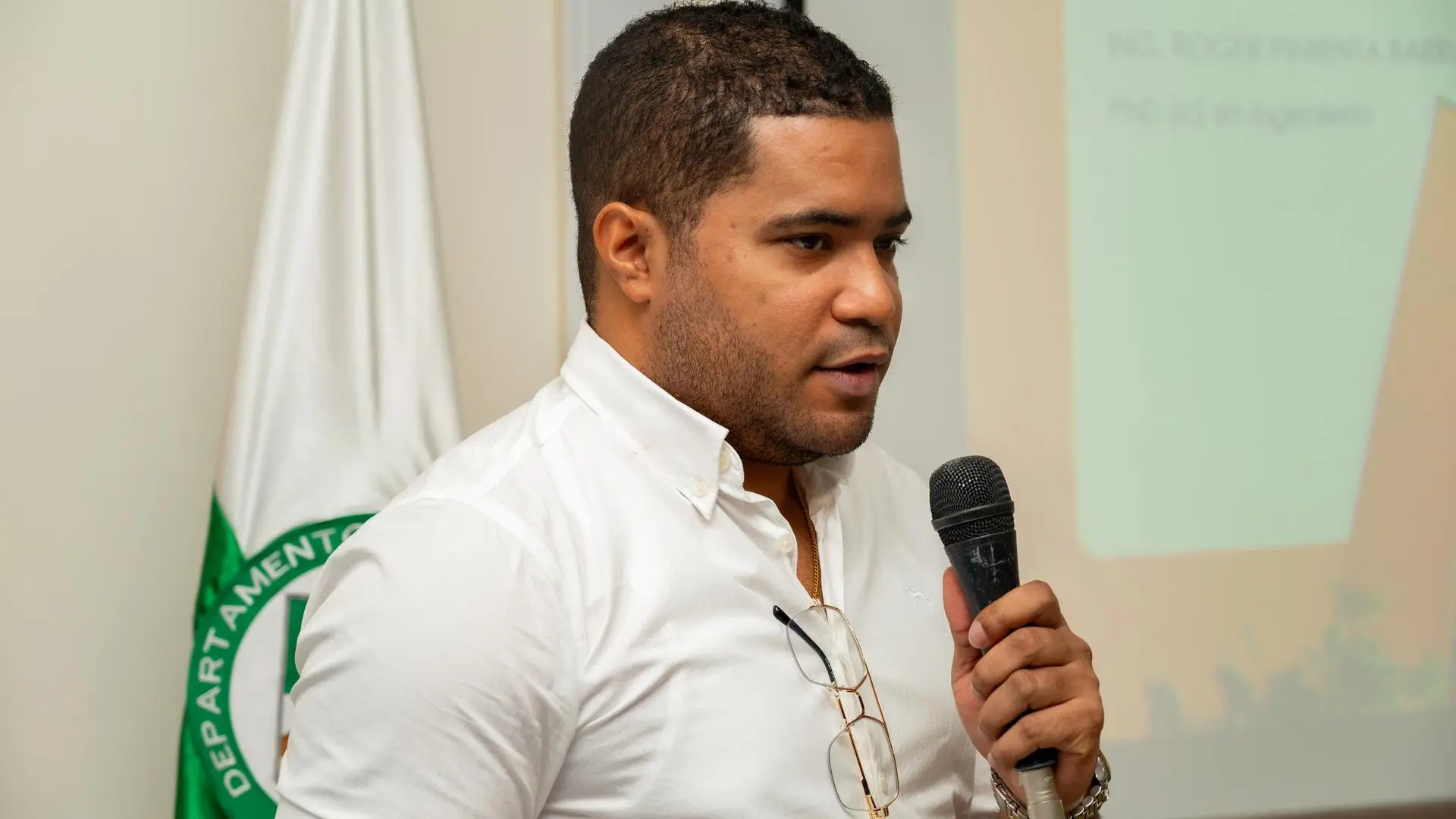 Hombre joven con camisa blanca sosteniendo un micrófono mientras habla durante una presentación. Lleva gafas colgadas en la camisa y al fondo se observa una bandera institucional y una proyección con texto parcialmente visible.