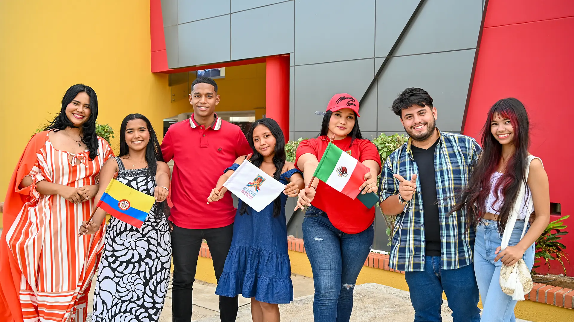 Grupo de jóvenes sonrientes sostiene banderas de Colombia, México y de la Universidad de La Guajira, frente a un edificio colorido, representando la diversidad y el intercambio académico.