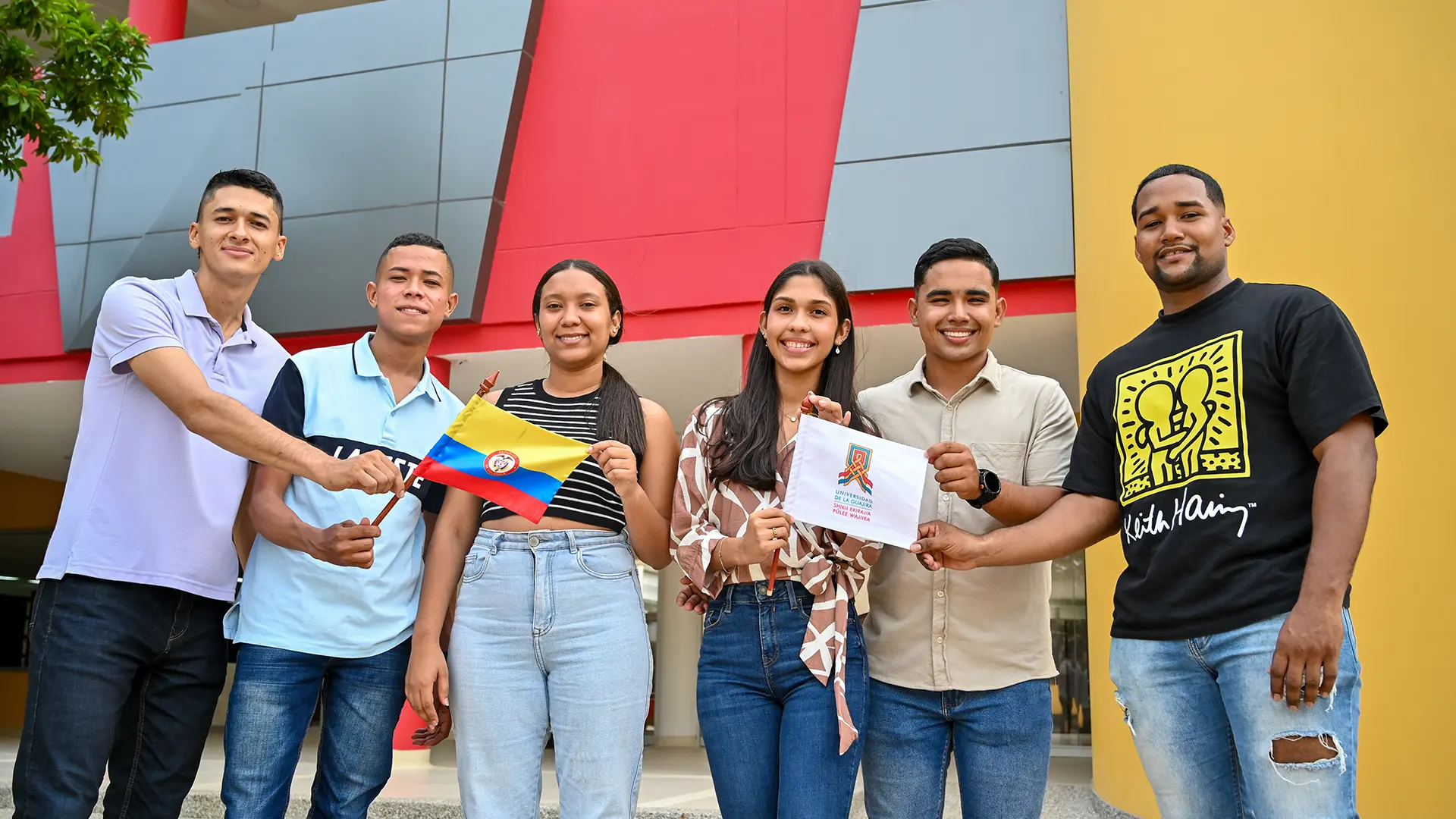 Grupo de jóvenes frente a un edificio moderno sostiene banderas de Colombia y de la Universidad de La Guajira, mostrando entusiasmo por la cooperación académica y la movilidad estudiantil.