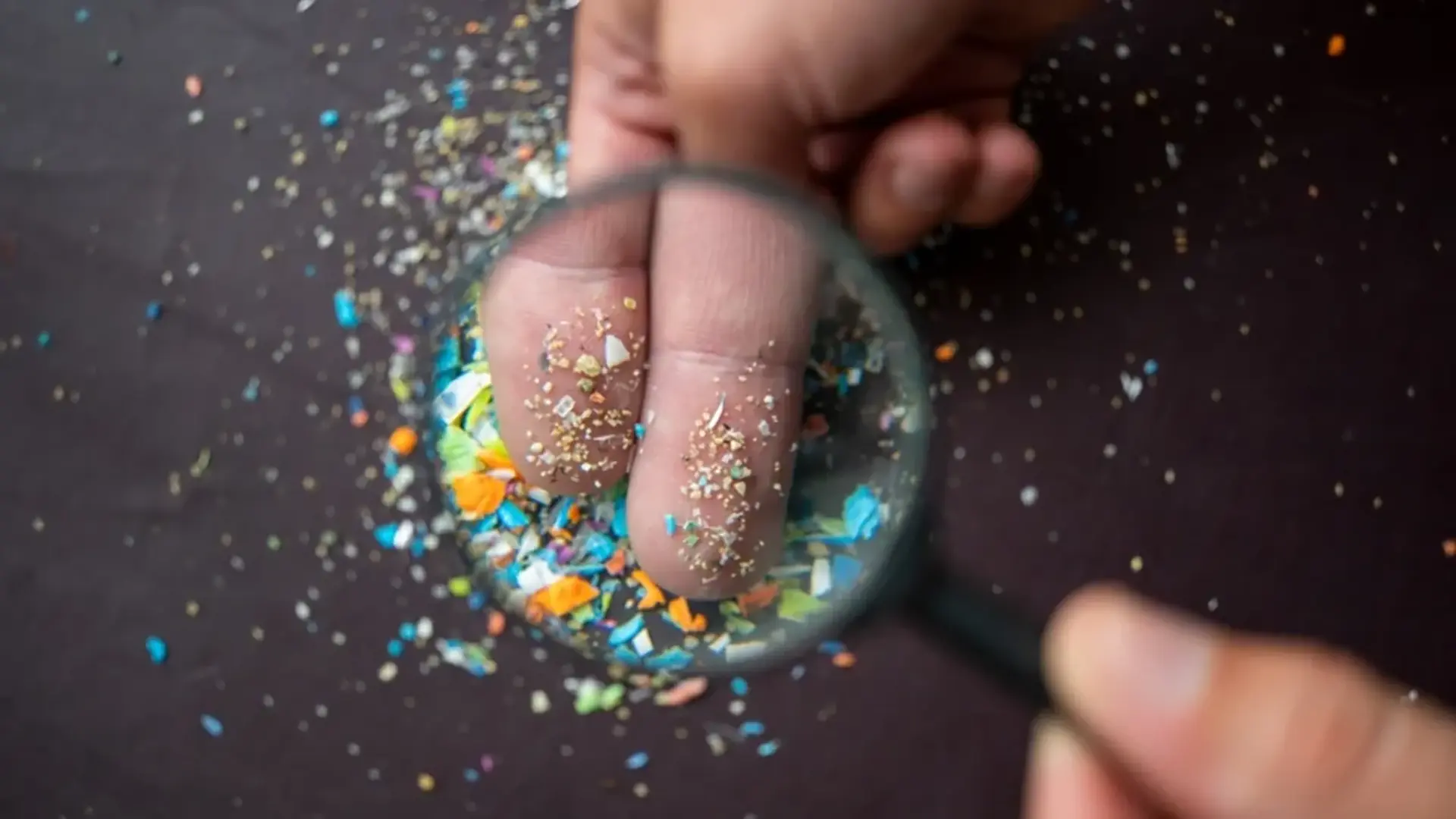 Dedos humanos cubiertos con partículas de microplásticos de colores, observados a través de una lupa sobre una superficie oscura.