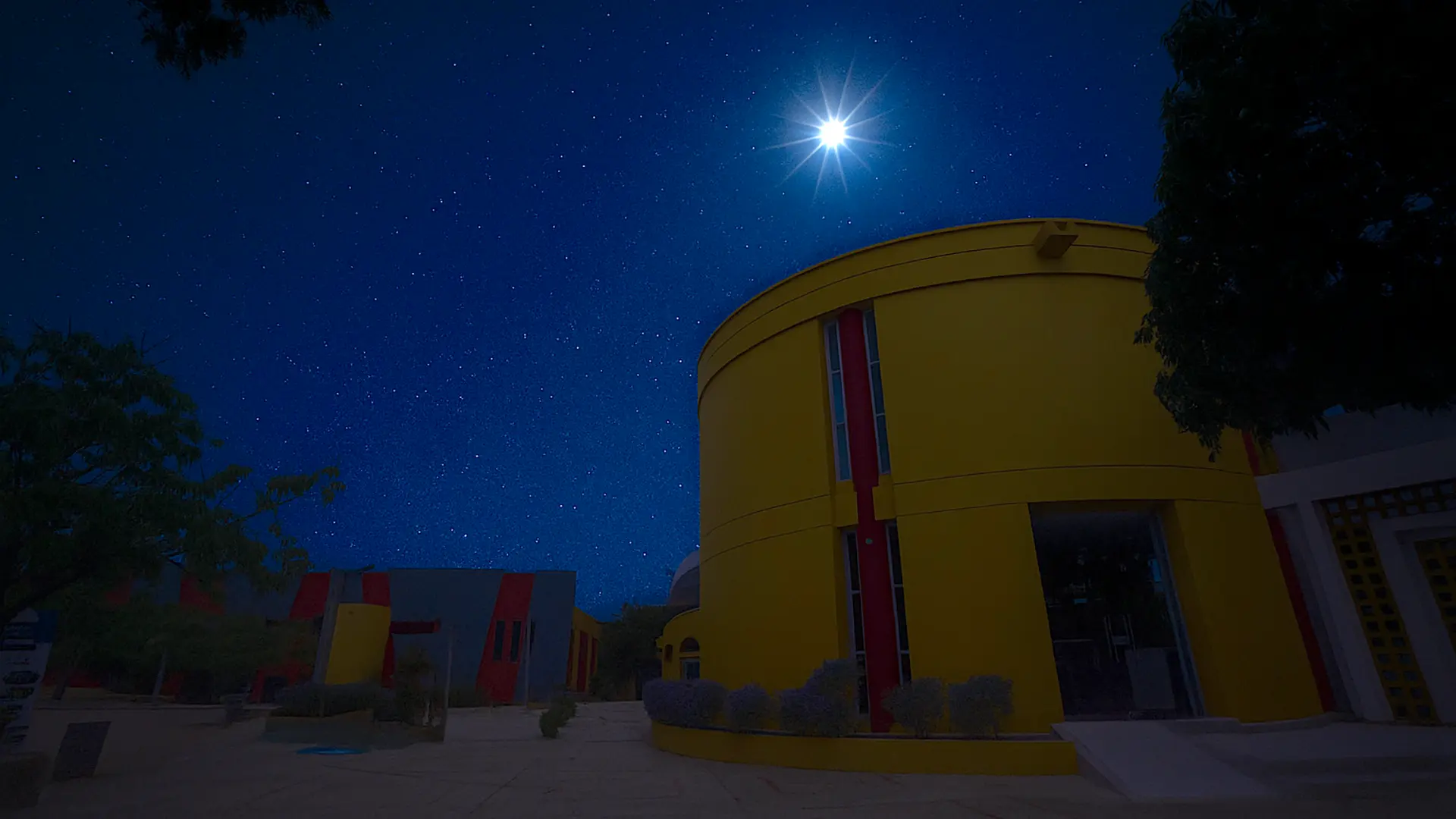 Vista nocturna del edificio redondo de color amarillo con detalles rojos del campus de la Universidad de La Guajira, bajo un cielo estrellado iluminado por una luna brillante. La escena transmite tranquilidad y destaca la arquitectura colorida del lugar en contraste con el cielo azul profundo.