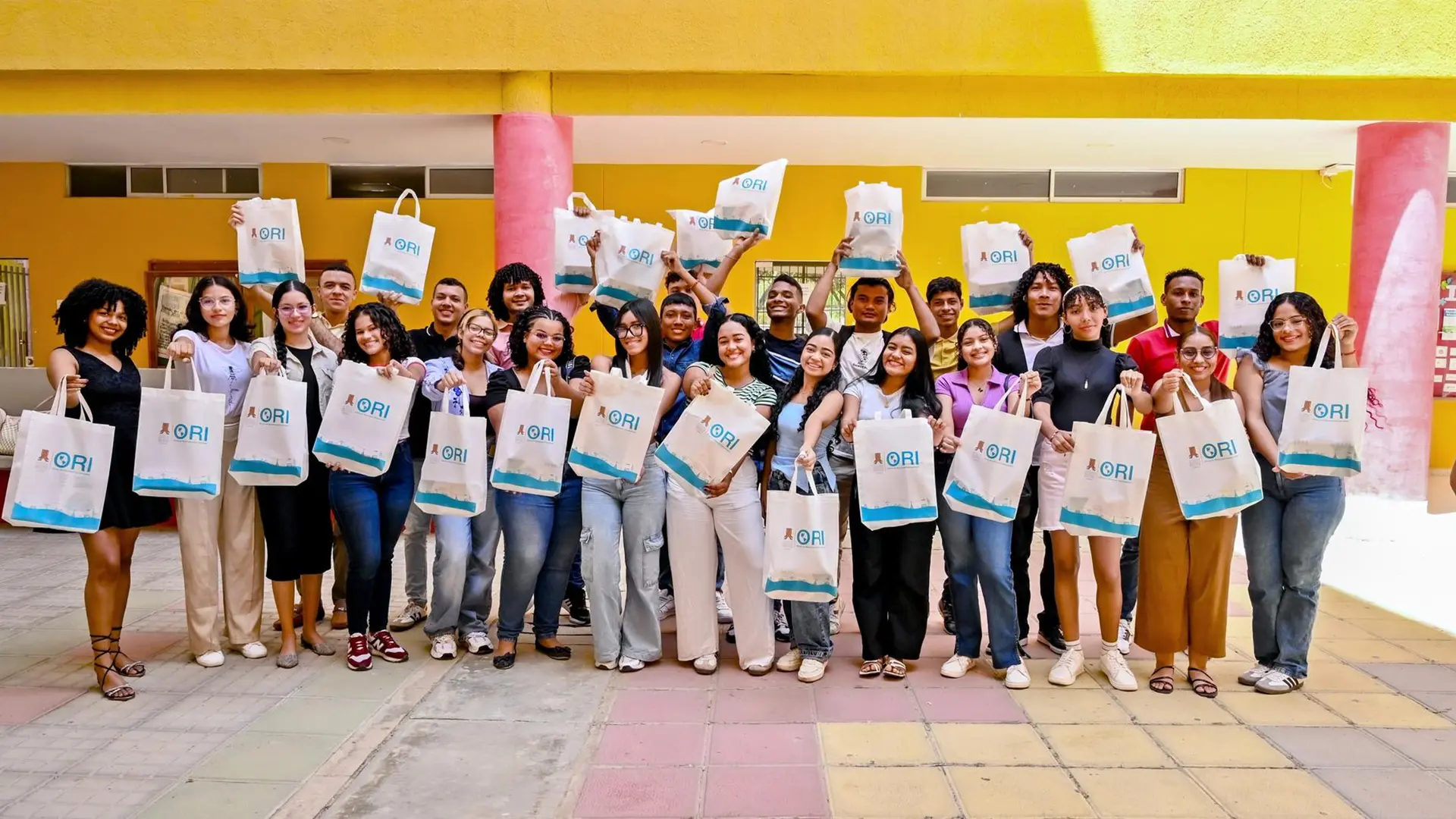 Grupo de estudiantes jóvenes, diversos y sonrientes posan en un patio con paredes amarillas y columnas rosadas, sosteniendo bolsas ecológicas blancas con el logo de la Oficina de Relaciones Internacionales (ORI).
