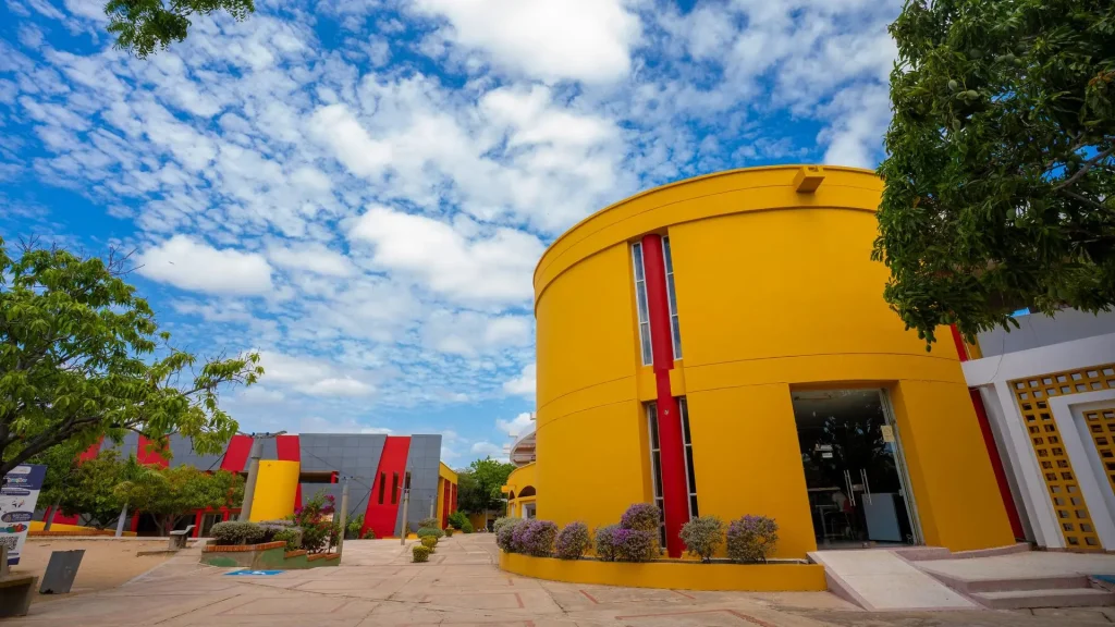Vista diurna de una moderna estructura universitaria con fachada amarilla y detalles en rojo, rodeada de árboles y cielo azul con nubes esponjosas.