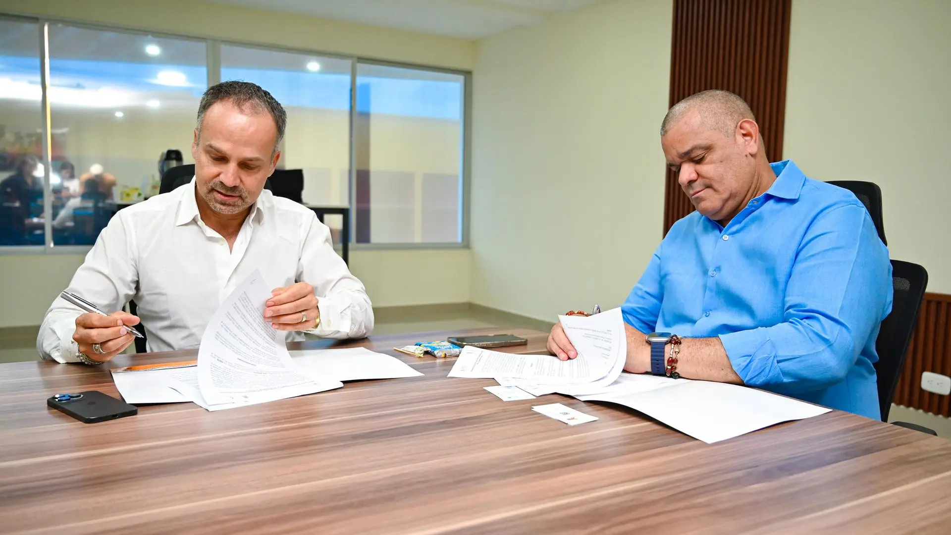 Dos hombres sentados en una mesa de reuniones firman documentos oficiales. Ambos visten camisas formales, uno blanca y el otro azul, y revisan cuidadosamente los papeles frente a ellos. El entorno es una oficina moderna con ventanales de fondo.