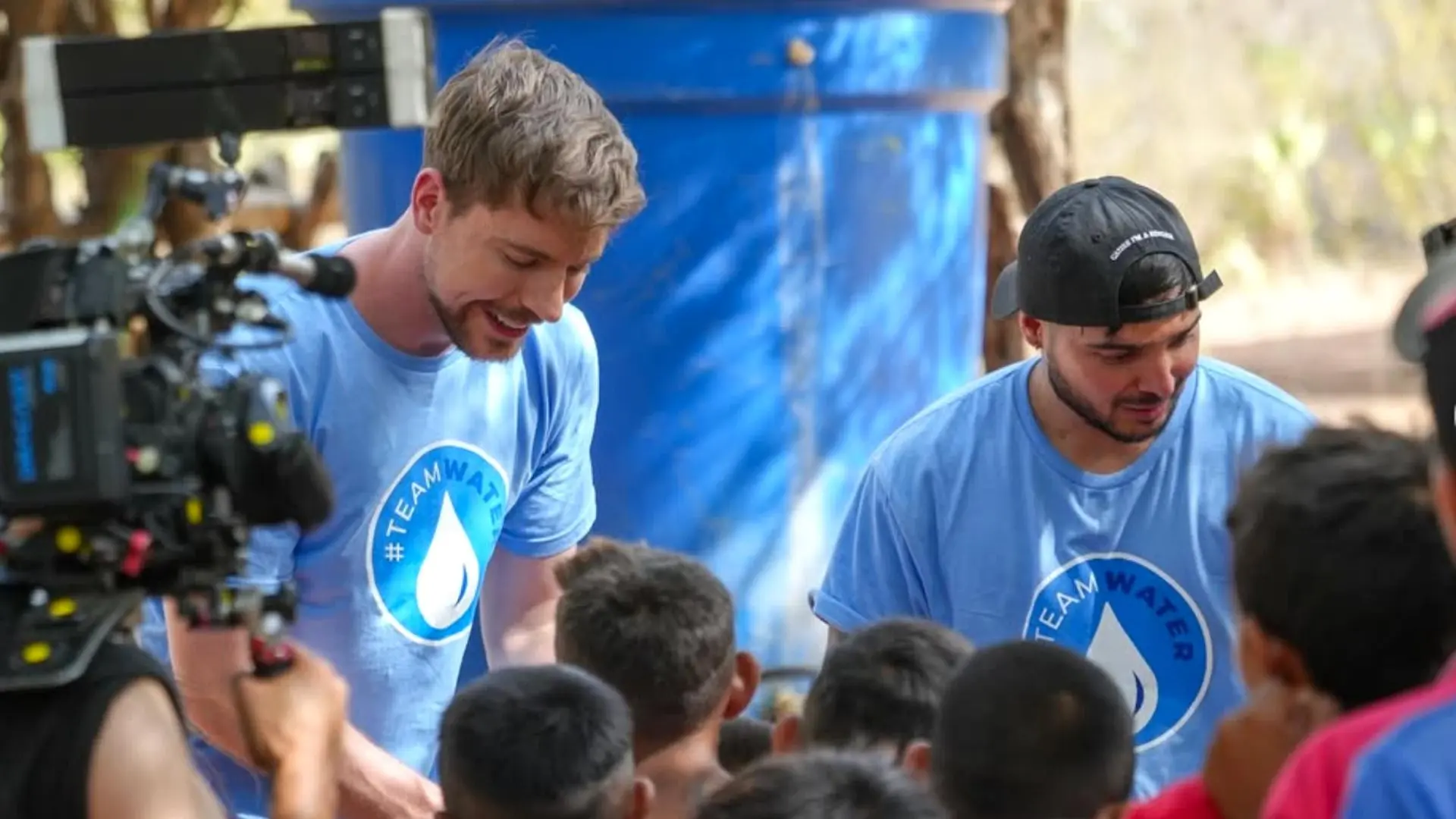 Dos hombres con camisetas azules de "#TEAMWATER" interactúan con un grupo de niños en una comunidad. Uno de ellos habla mientras el otro observa atentamente. A la izquierda, una cámara graba la escena. De fondo, se ve un tanque azul y vegetación árida.