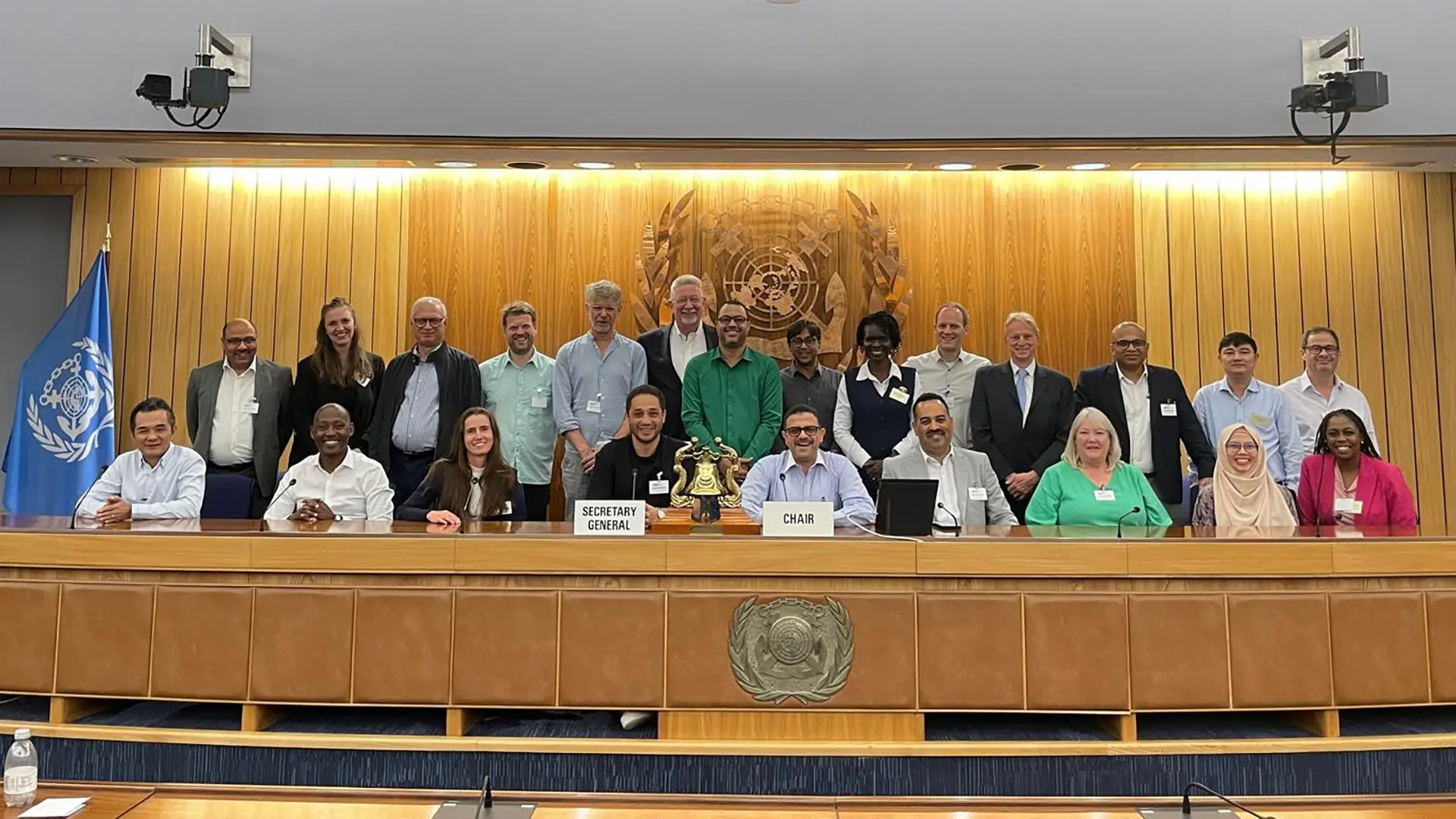 Un grupo diverso de personas posando para una foto oficial en una sala de conferencias de la Organización Marítima Internacional (OMI). En la mesa principal hay carteles que dicen "Secretary General" y "Chair", con un símbolo institucional entre ellos. Detrás del grupo se observa el emblema de la OMI y una bandera azul con el logo de la organización.