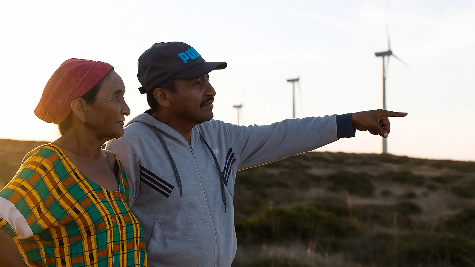 Una mujer indígena con vestido colorido y un hombre con gorra y sudadera observan el horizonte mientras el hombre señala hacia una fila de aerogeneradores eólicos, destacando la convivencia entre comunidades locales y energías renovables.