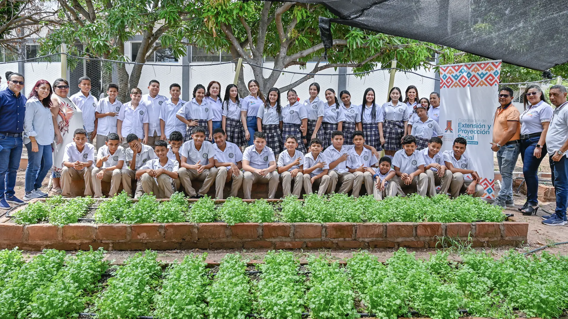 Docentes, directivos y estudiantes de secundaria posan en un huerto escolar sembrado de cilantro. Al fondo, bajo la sombra de árboles y mallas, se encuentra un cartel de la Coordinación de Extensión y Proyección Social – Sede Villanueva, que identifica la actividad comunitaria.