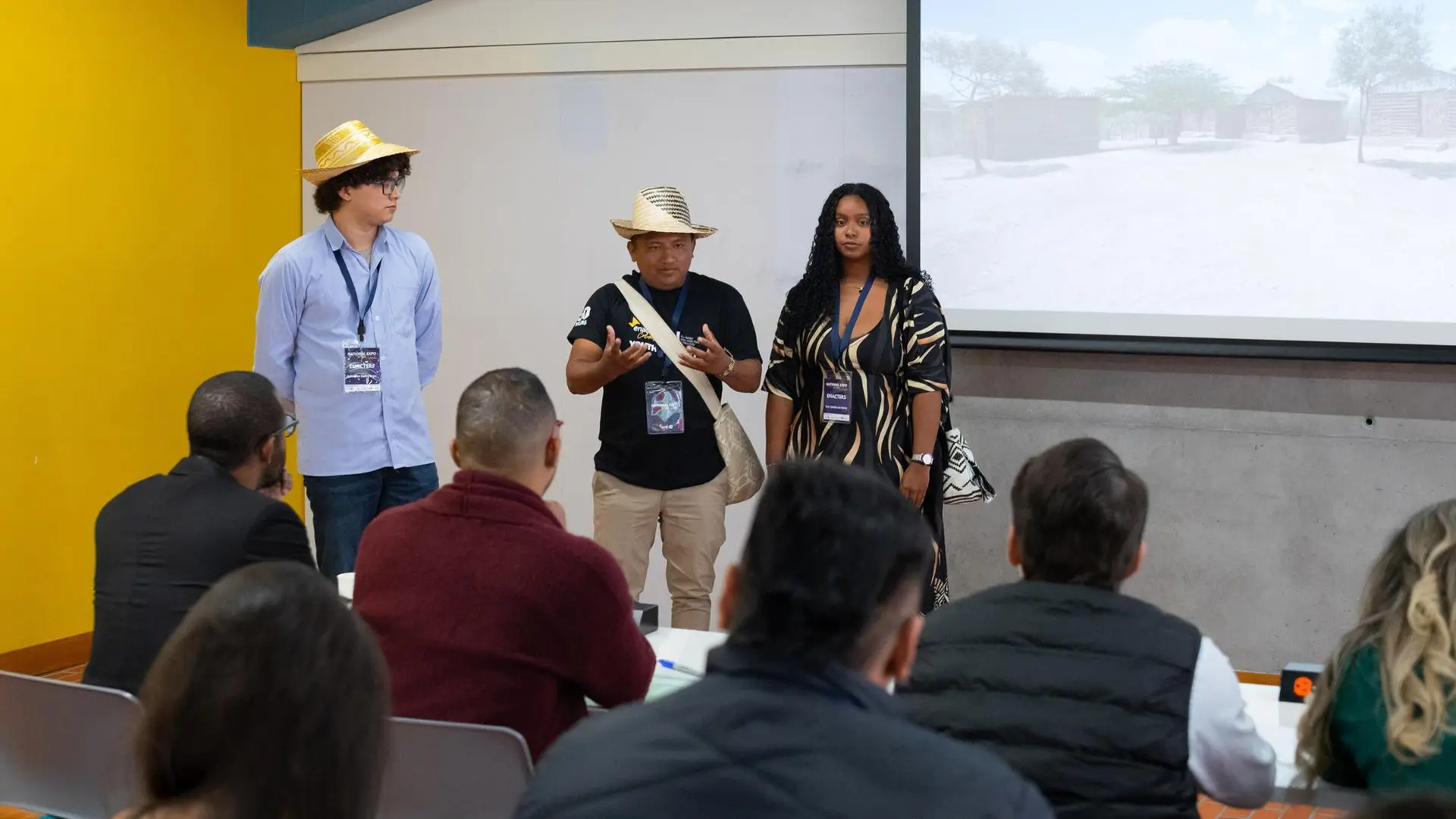 Tres jóvenes (dos hombres y una mujer) están exponiendo frente a un jurado o público. Uno de ellos utiliza sombrero vueltiao y hace gestos con las manos mientras habla, lo que indica que está explicando o defendiendo una propuesta. Al fondo, se proyecta una imagen de un entorno rural, posiblemente relacionado con la temática de su proyecto.