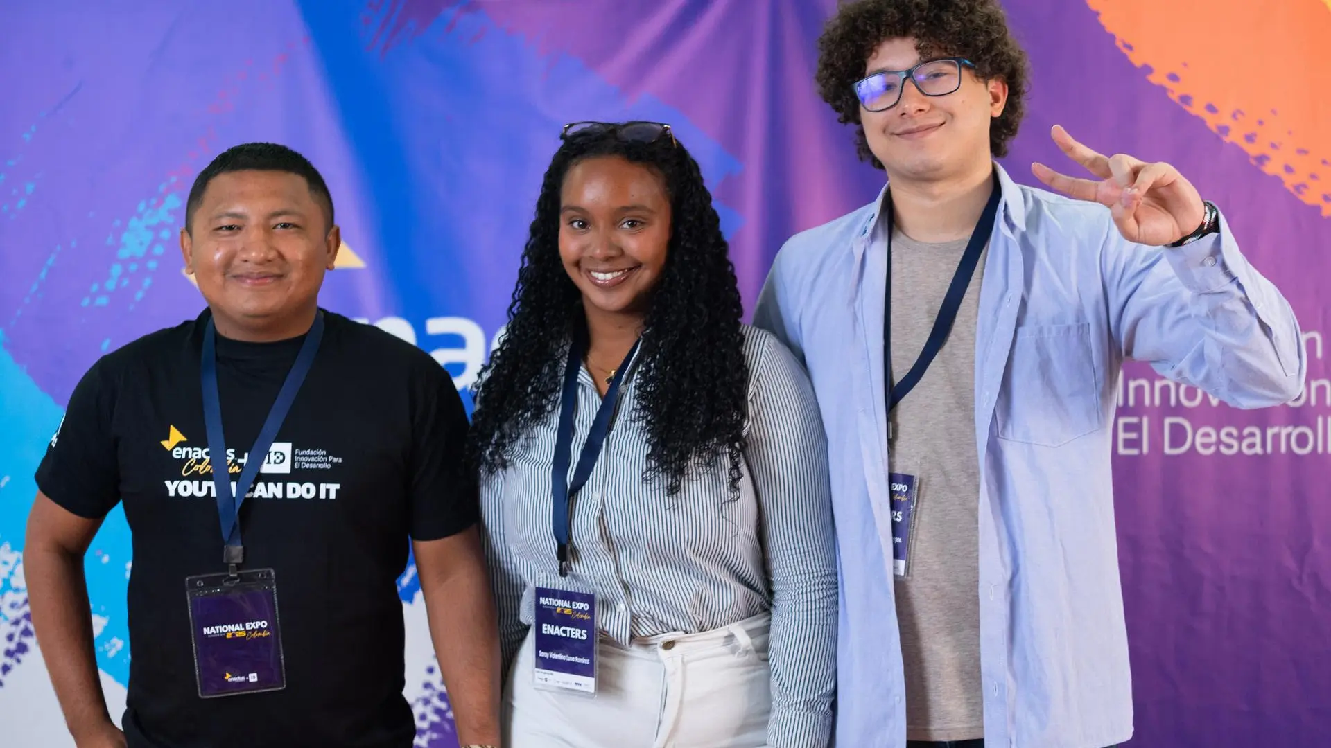 Tres jóvenes posan sonrientes frente a un backdrop oficial del evento que tiene los logos de Enactus Colombia y mensajes de innovación. Llevan gafetes de acreditación con la palabra “ENACTERS” y el texto National Expo Colombia, lo que confirma que participaron en la Expo Nacional Enactus, una competencia de impacto social y empresarial.