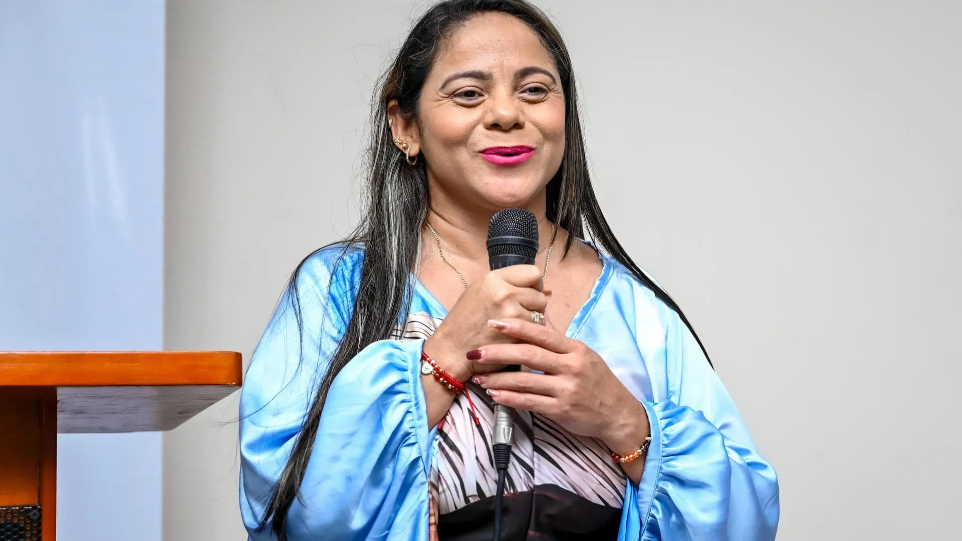 Una mujer sonriente, vestida con una capa azul satinada, sostiene un micrófono mientras se dirige al público en un evento institucional en Uniguajira.