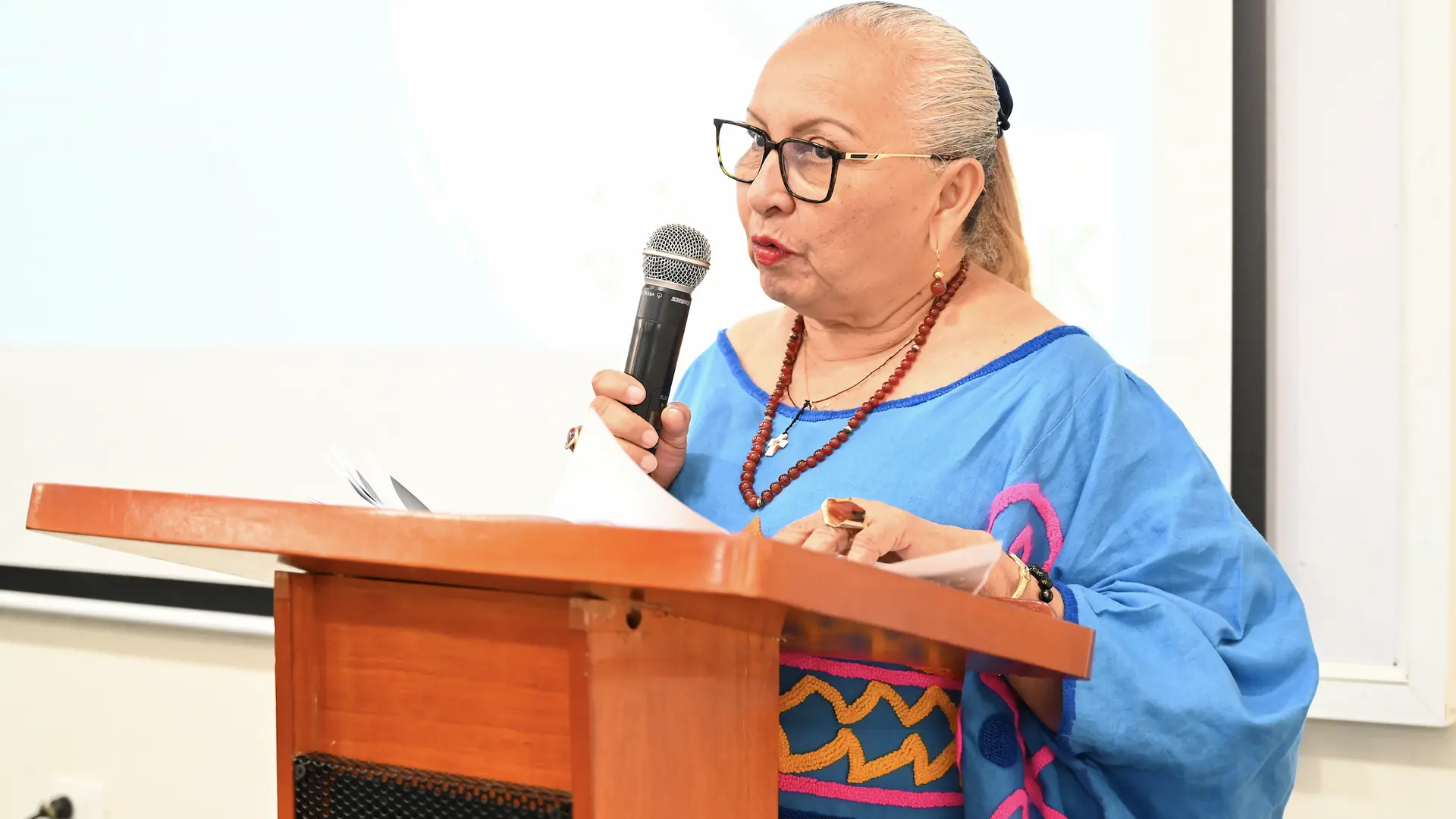 Una mujer de cabello recogido y gafas, vestida con un atuendo azul tradicional, habla en un podio con micrófono durante un evento académico o cultural en Uniguajira.