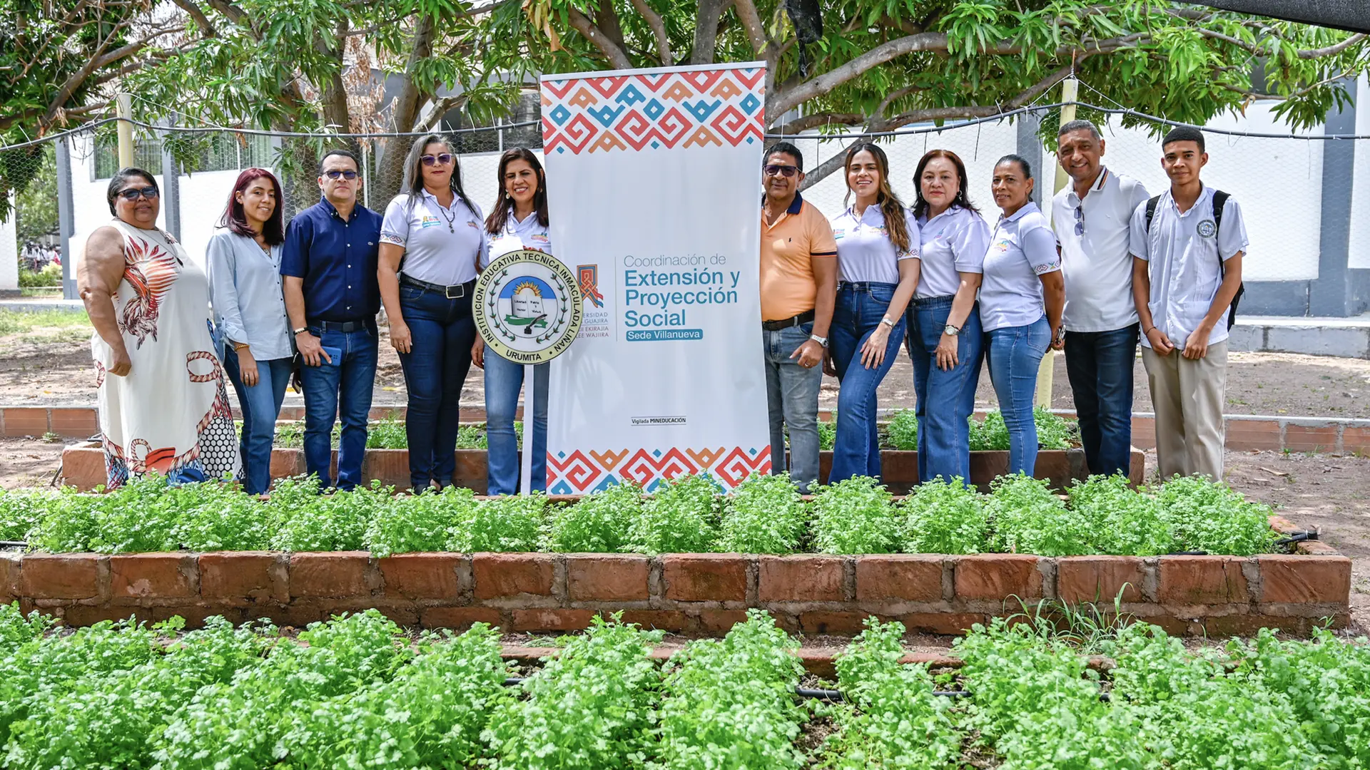 Un grupo de docentes y representantes institucionales posan de pie frente a un huerto escolar de cilantro. En el centro se observa un cartel de la Coordinación de Extensión y Proyección Social – Sede Villanueva, acompañado del logo de la institución educativa.