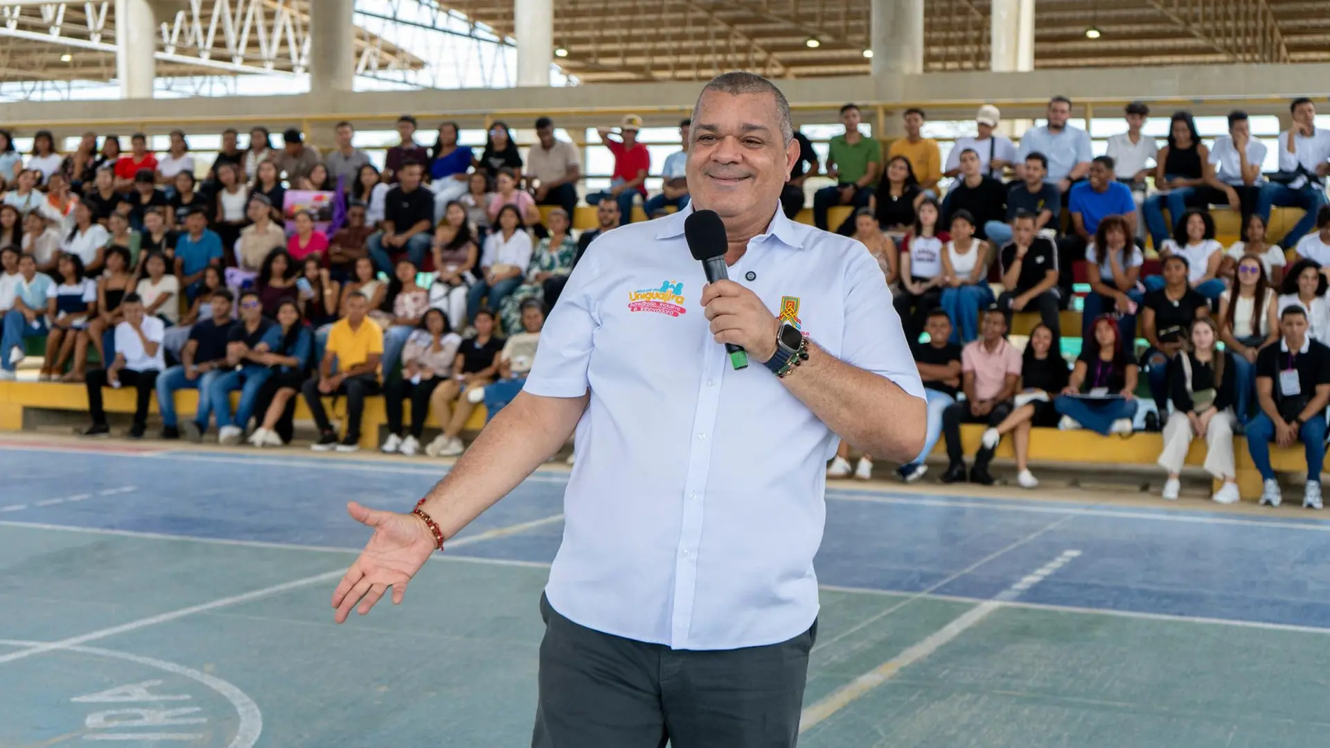 Hombre con micrófono en mano hablando frente a una audiencia en un coliseo cubierto. Viste camisa blanca con logos coloridos bordados relacionados con un evento universitario. Al fondo, se observa a un grupo numeroso de estudiantes sentados en las gradas.