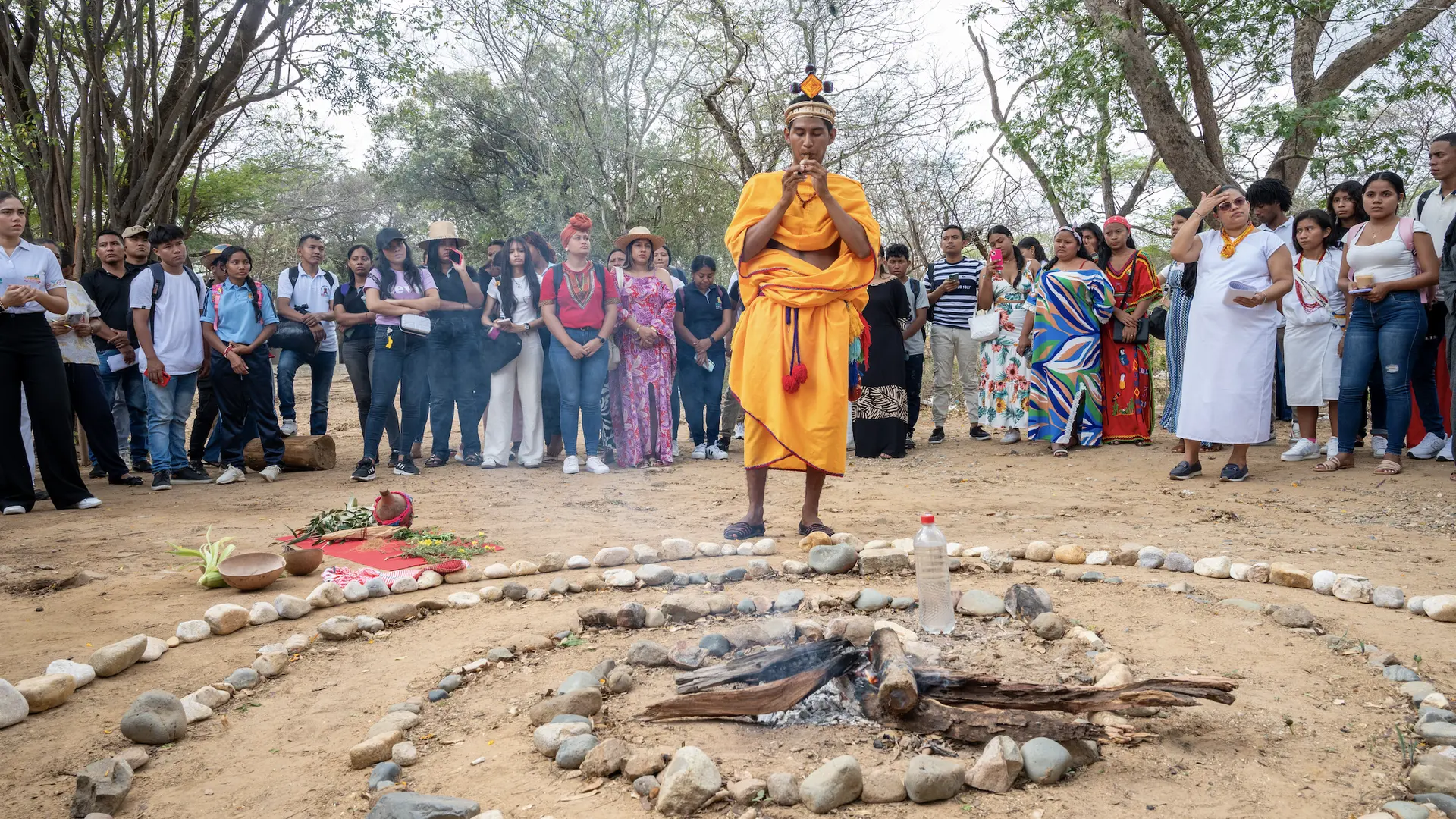 Un grupo de estudiantes y docentes de Uniguajira observan una ceremonia cultural indígena en un espacio abierto, donde un hombre vestido con atuendo tradicional naranja realiza un ritual alrededor de un círculo de piedras y fuego.