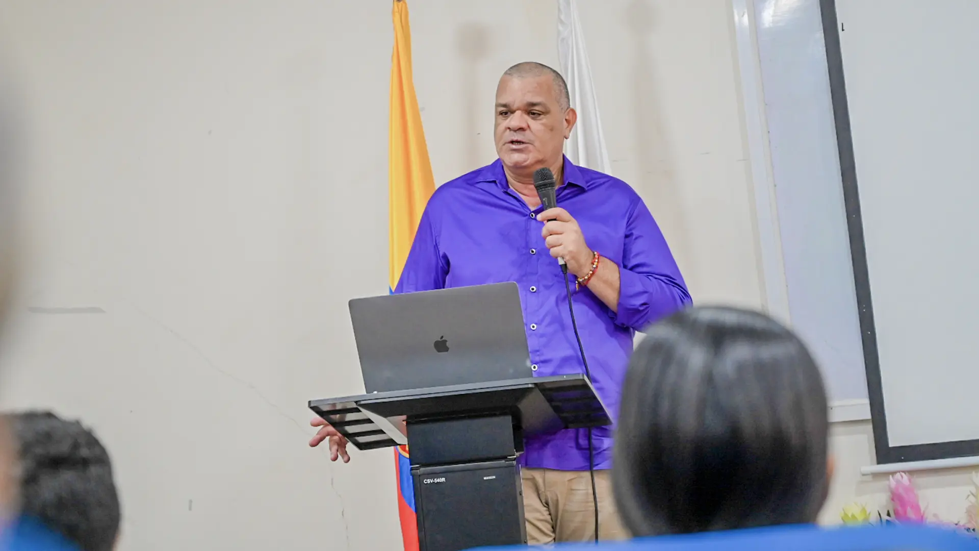 Hombre de pie frente a un atril con un computador portátil, hablando con micrófono en mano. Al fondo se observan las banderas de Colombia y de una institución.