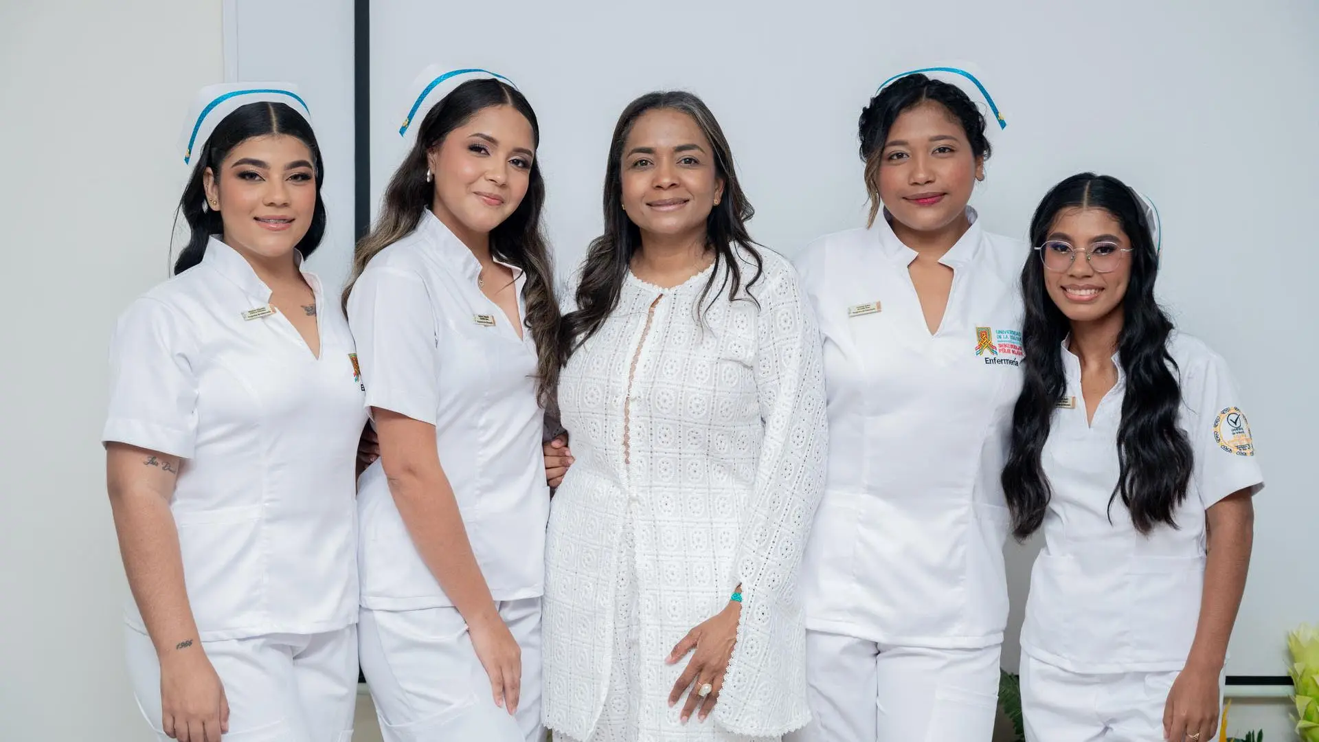 Grupo de cuatro estudiantes de enfermería con uniforme blanco y cofias posan sonrientes junto a una mujer vestida de blanco, en un ambiente formal de ceremonia.