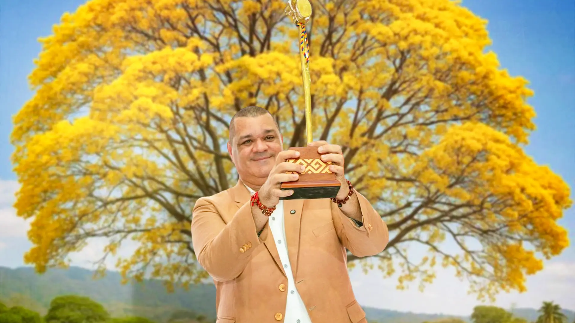 Hombre sonriente sostiene un trofeo dorado con base decorada en tonos marrón y dorado. Viste saco beige y camisa blanca. Al fondo se aprecia un gran árbol de flores amarillas que cubre casi toda la escena.
