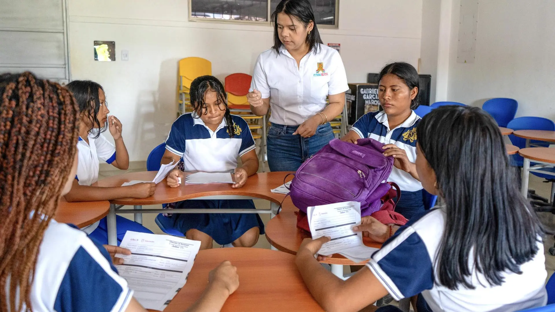 Grupo de estudiantes en uniforme escolar azul y blanco, sentados en mesas circulares dentro de un salón de clases, reciben orientación de una docente que les entrega indicaciones mientras revisan documentos.