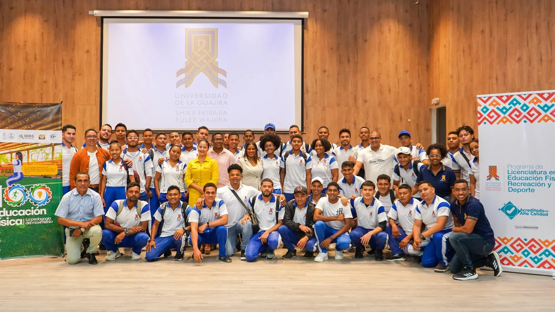 Grupo de estudiantes y docentes del programa de Licenciatura en Educación Física, Recreación y Deporte de la Universidad de La Guajira posan en un auditorio, acompañados de afiches institucionales y con el logo de la universidad proyectado en pantalla.