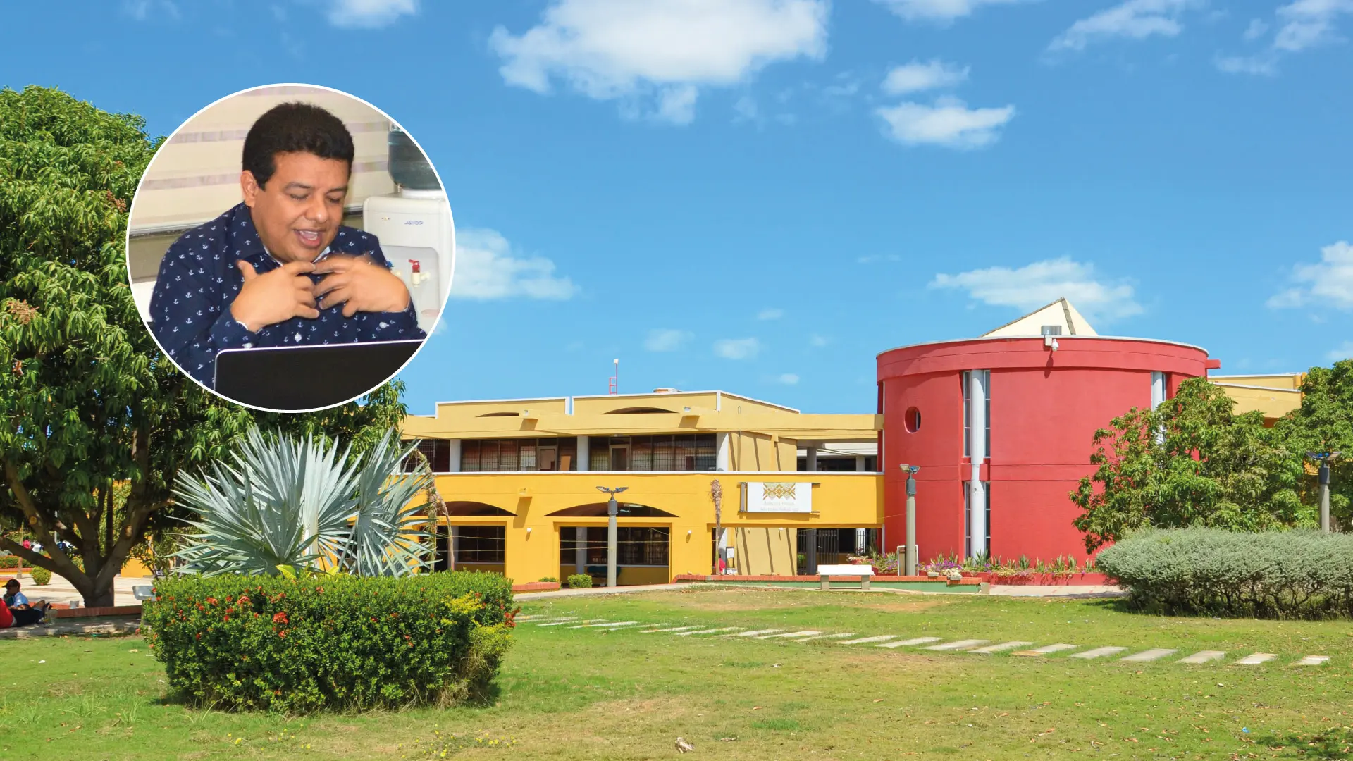 Vista del campus de la Universidad de La Guajira en un día soleado, con edificios de colores amarillo y rojo rodeados de jardines. En un recuadro circular se muestra a un participante hablando frente a un computador durante una reunión virtual.