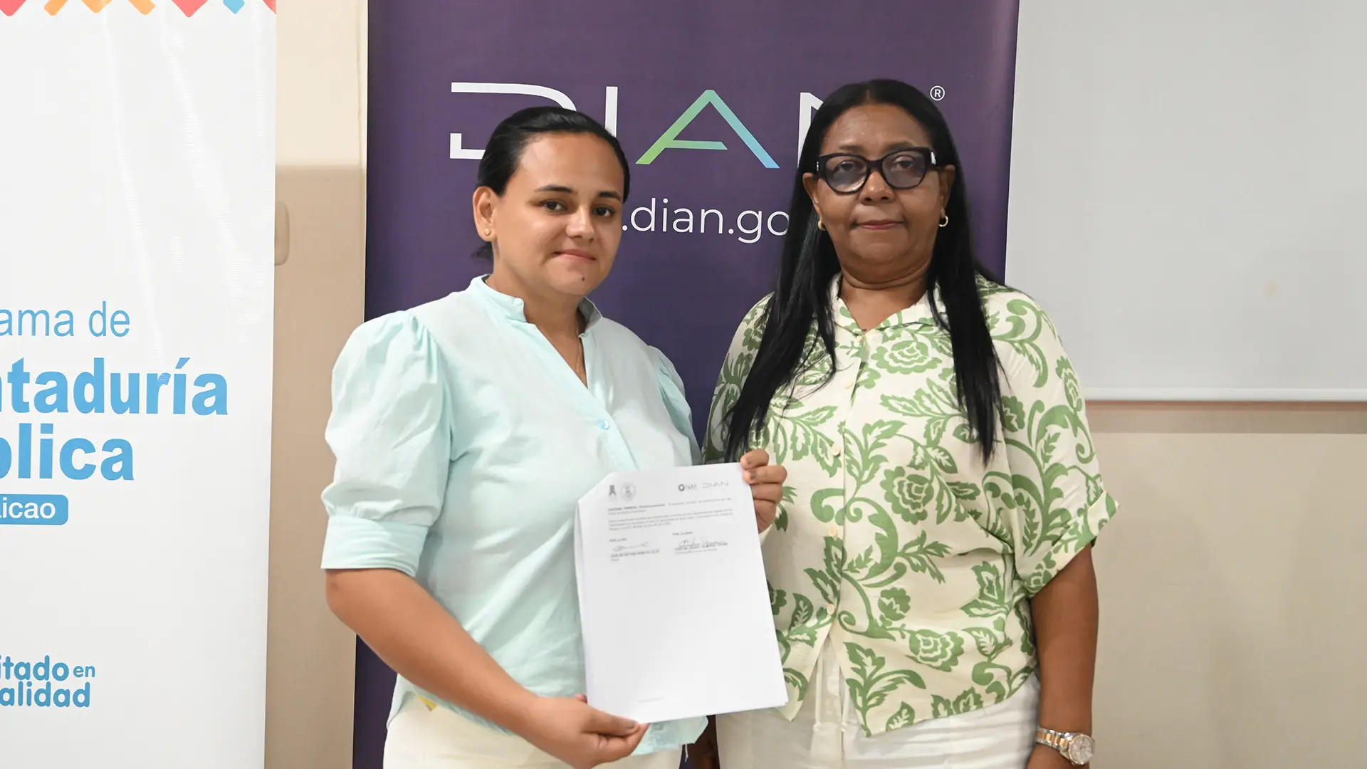 Dos mujeres posan frente a pendones institucionales; una de ellas sostiene un documento oficial de la DIAN. Ambas visten ropa clara y miran a la cámara en un ambiente académico o administrativo.
