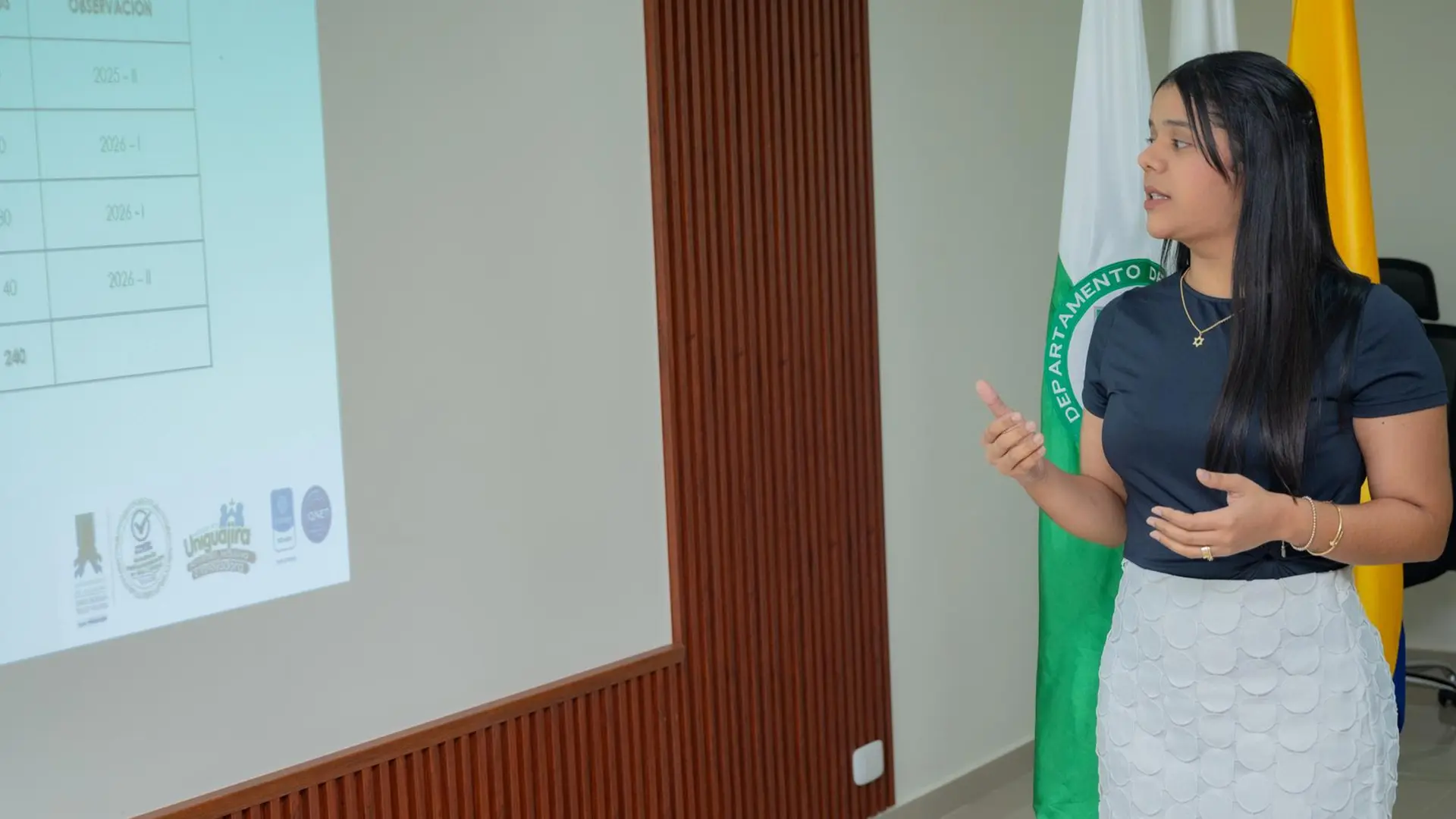 Una mujer joven realiza una presentación frente a una proyección que muestra un cuadro con información de cupos y observaciones académicas. Ella gesticula mientras explica, y detrás se observan las banderas de Colombia y del departamento de La Guajira. La escena refleja un contexto académico o institucional.