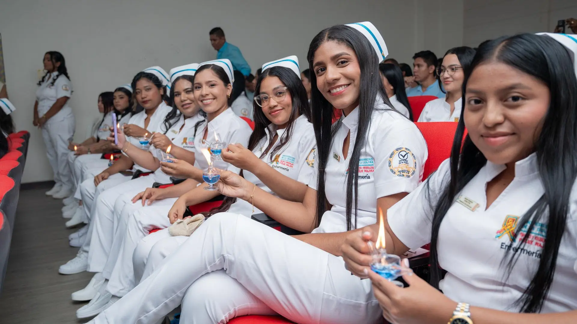 Estudiantes de enfermería sentados en un auditorio sostienen lámparas encendidas en sus manos, como parte de un ritual simbólico de la profesión.