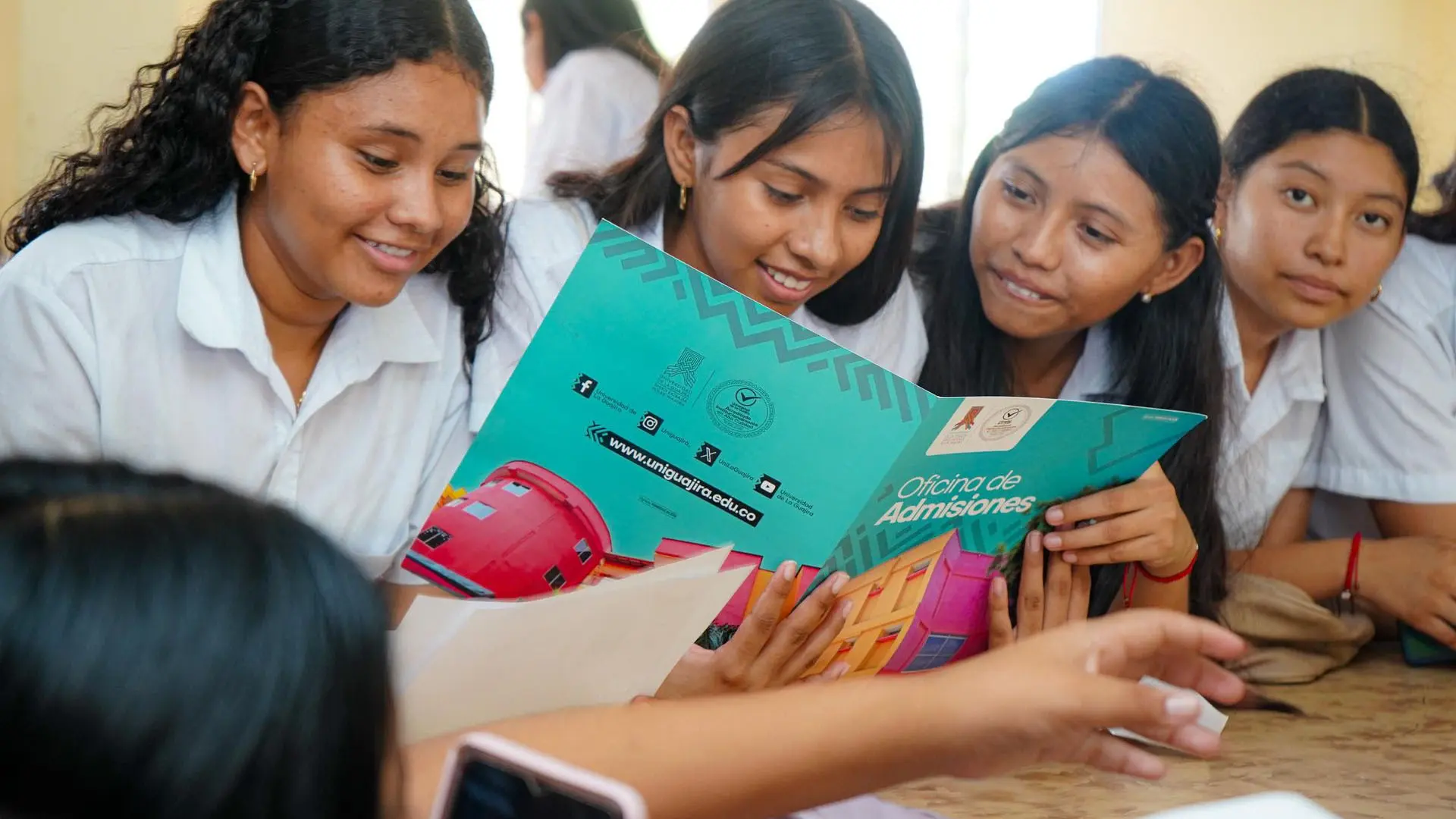 Varías jóvenes estudiantes sonríen y observan juntas un folleto colorido de la Oficina de Admisiones de la Universidad de La Guajira, sentadas alrededor de una mesa en un aula.