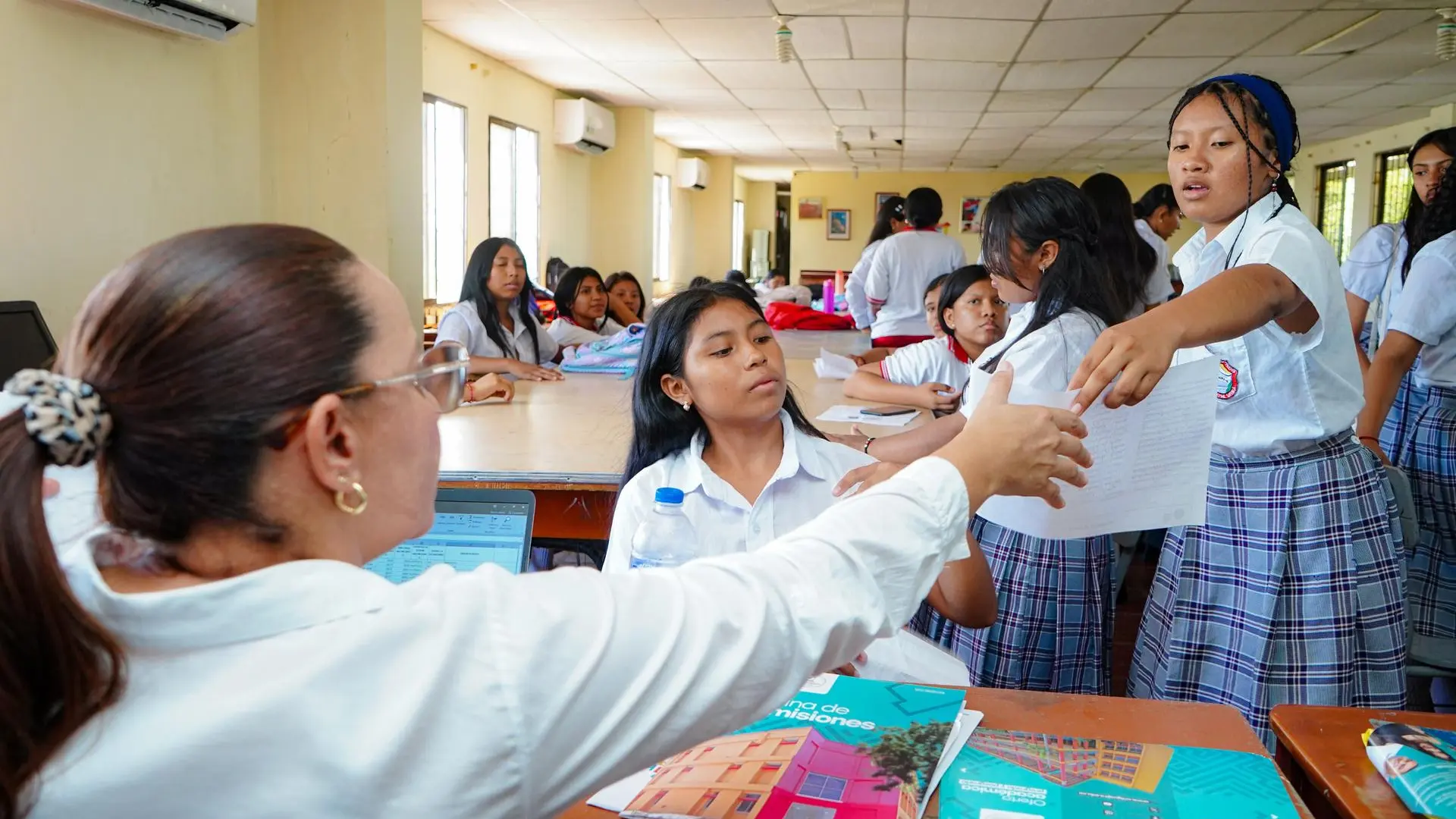 En un salón de clases, varias estudiantes con uniforme escolar de blusa blanca y falda a cuadros participan en una actividad académica. Una de ellas entrega una hoja a la docente, mientras otras observan y esperan su turno alrededor de una mesa larga. La escena refleja un ambiente educativo y de interacción en grupo.