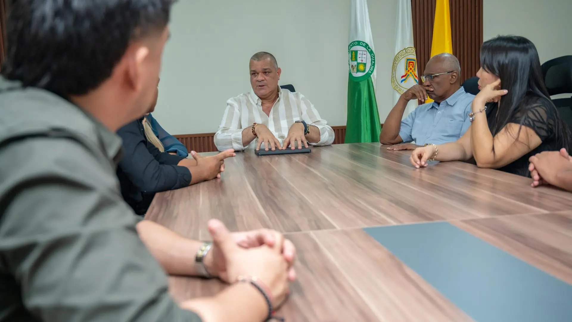 Un grupo de personas participa en una reunión alrededor de una mesa de madera. En el centro, un hombre con camisa blanca de rayas habla mientras apoya sus manos sobre una carpeta. A su alrededor, los demás asistentes escuchan con atención. Al fondo se observan las banderas de Colombia, del Departamento de La Guajira.