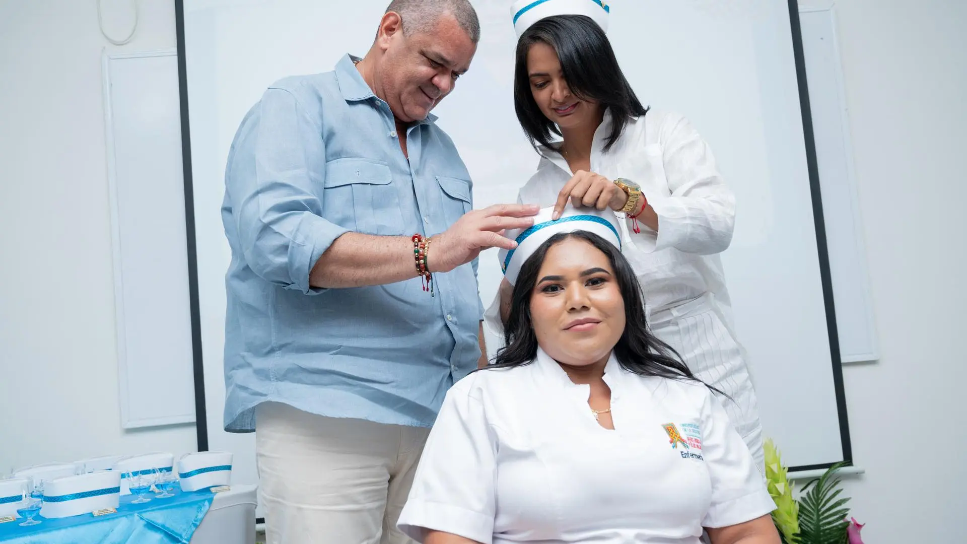 Estudiante de enfermería sentada recibe la colocación de la cofia blanca con ribete azul por parte de dos personas, durante una ceremonia simbólica de imposición de cofias.