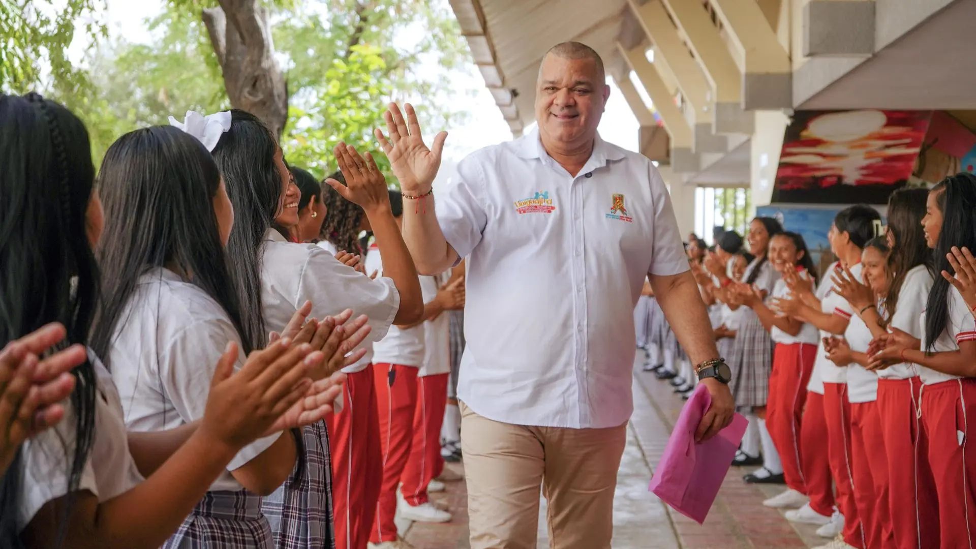 Un hombre adulto con camisa blanca institucional camina sonriente por un pasillo escolar, mientras estudiantes uniformados forman un corredor aplaudiéndole y dándole la bienvenida con entusiasmo.