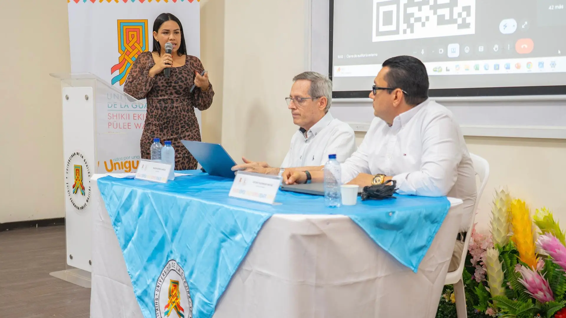 Una mujer de pie, con micrófono en mano, interviene en un auditorio frente a dos hombres sentados en una mesa cubierta con mantel azul y el logo de la Universidad de La Guajira. Al fondo se proyecta una presentación con un código QR