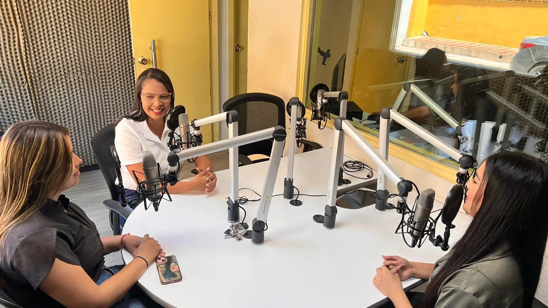 Tres mujeres participan en una conversación en una cabina de radio. Están sentadas alrededor de una mesa con micrófonos profesionales, sonriendo y conversando en un ambiente de grabación. Al fondo se observa una ventana de vidrio que separa el área de control del estudio.