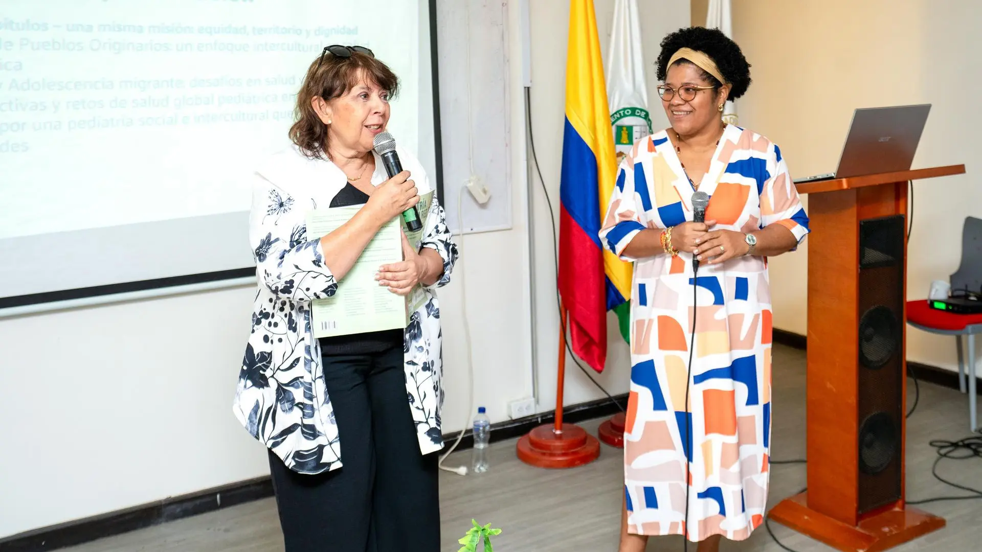“Dos mujeres durante una presentación académica en un auditorio; una sostiene un micrófono y un libro mientras la otra escucha y sonríe. Detrás de ellas se observan las banderas de Colombia y del Departamento de La Guajira.”