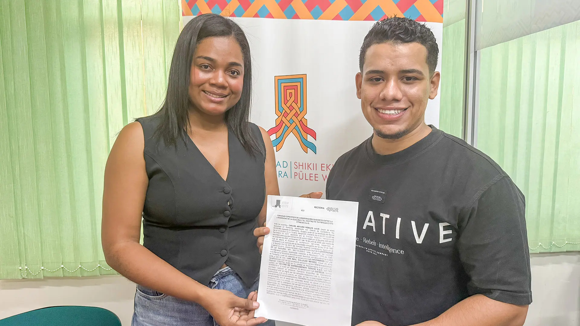 Dos personas sonríen mientras sostienen un documento. Una viste blusa negra y jeans, y la otra lleva una camiseta negra con letras blancas. En el fondo se distingue el logo de la Universidad de La Guajira, Sede Maicao, sobre un diseño con patrones geométricos de colores.
