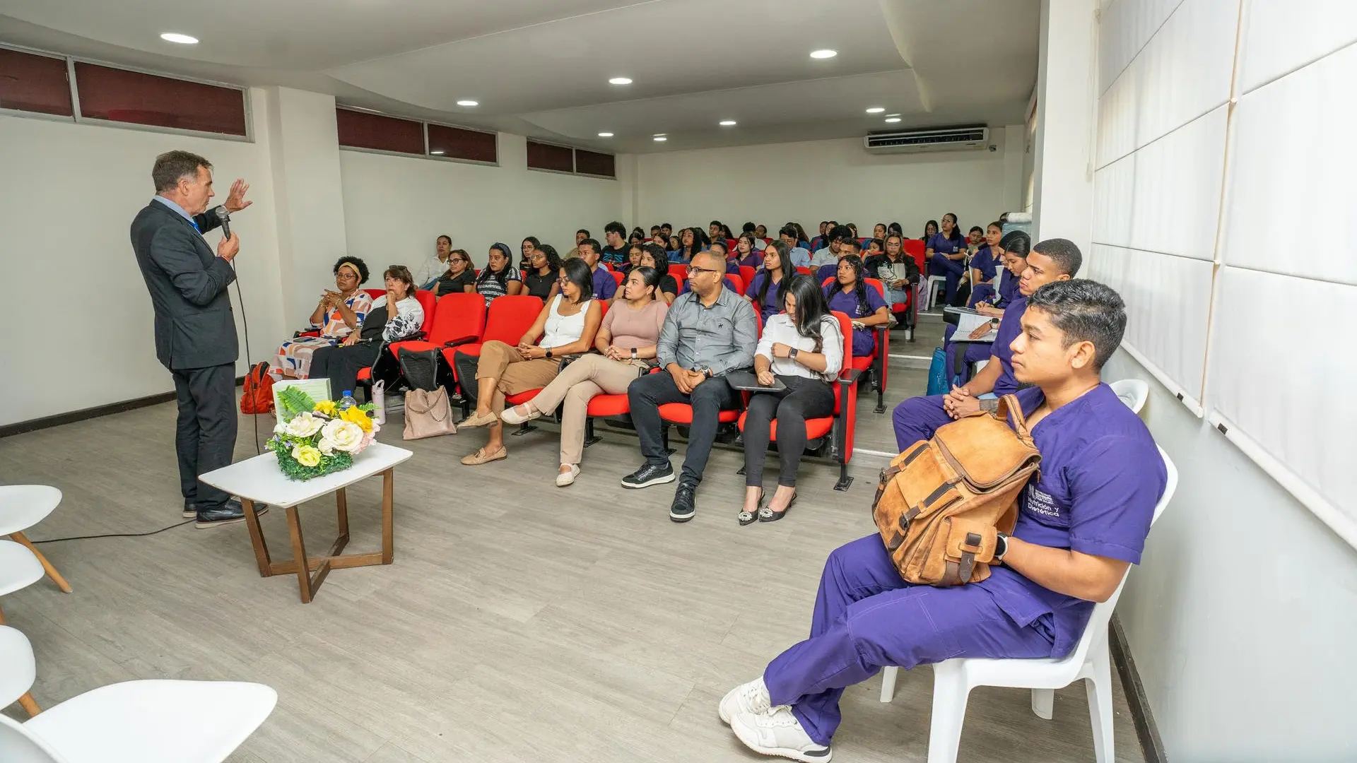 “Conferencia académica en un auditorio con asistentes sentados en sillas rojas. Un expositor de pie habla al público, mientras estudiantes y profesionales escuchan atentamente y toman notas.”