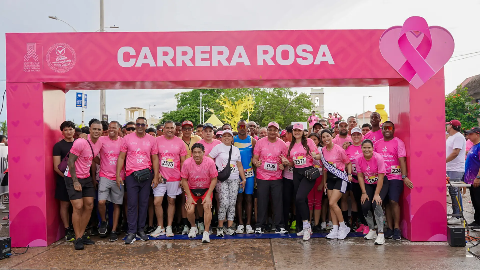 Grupo de mujeres y hombres con camisetas rosas levantando los brazos y aplaudiendo al aire, participando en el calentamiento previo a una carrera al aire libre.