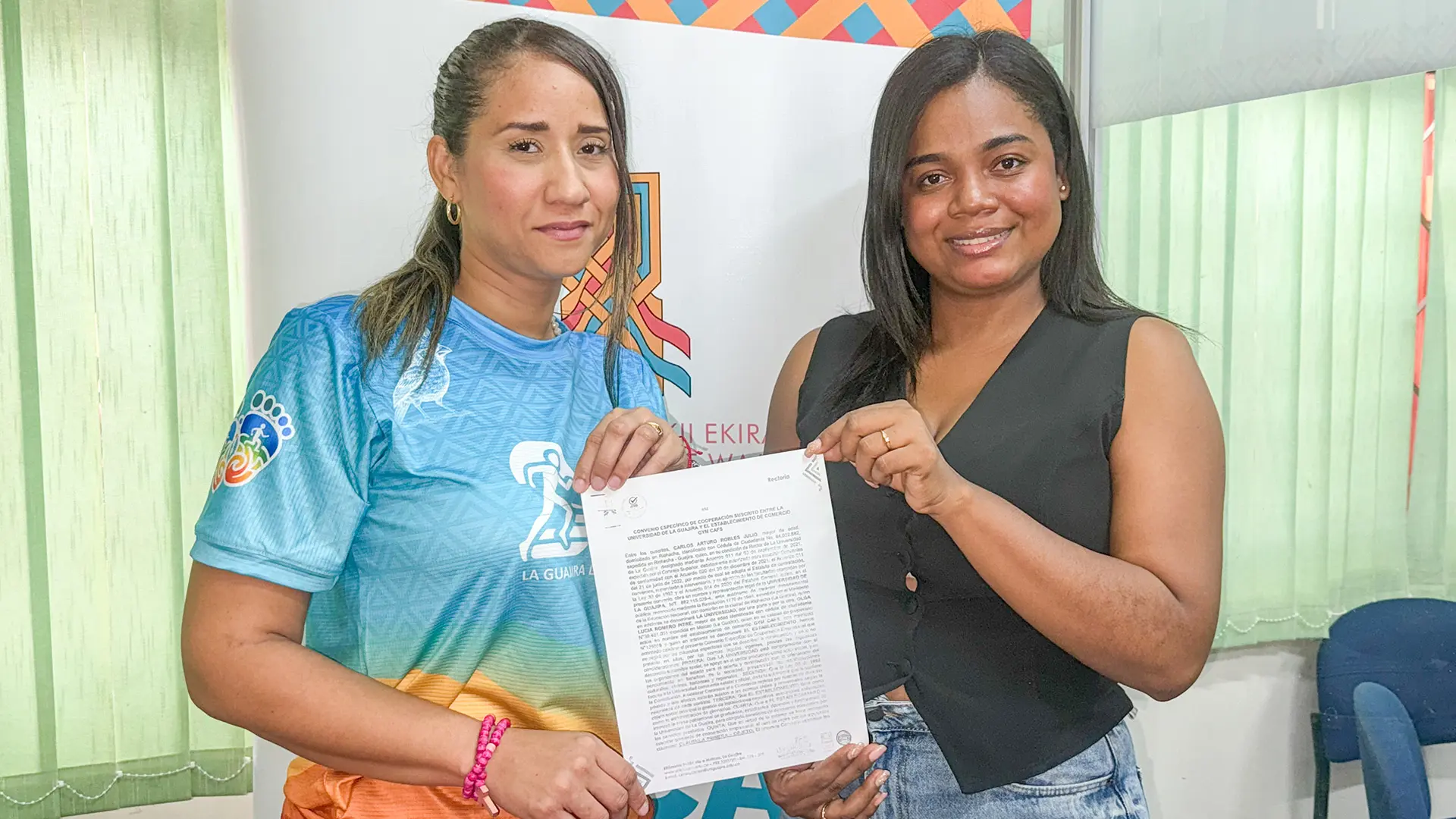 Dos personas posan sonriendo mientras sostienen un documento. Una de ellas viste una camiseta deportiva azul con detalles naranjas, y la otra usa un chaleco negro y jeans. Detrás se observa un cartel con el logo de la Universidad de La Guajira, Sede Maicao, y un diseño colorido de fondo.