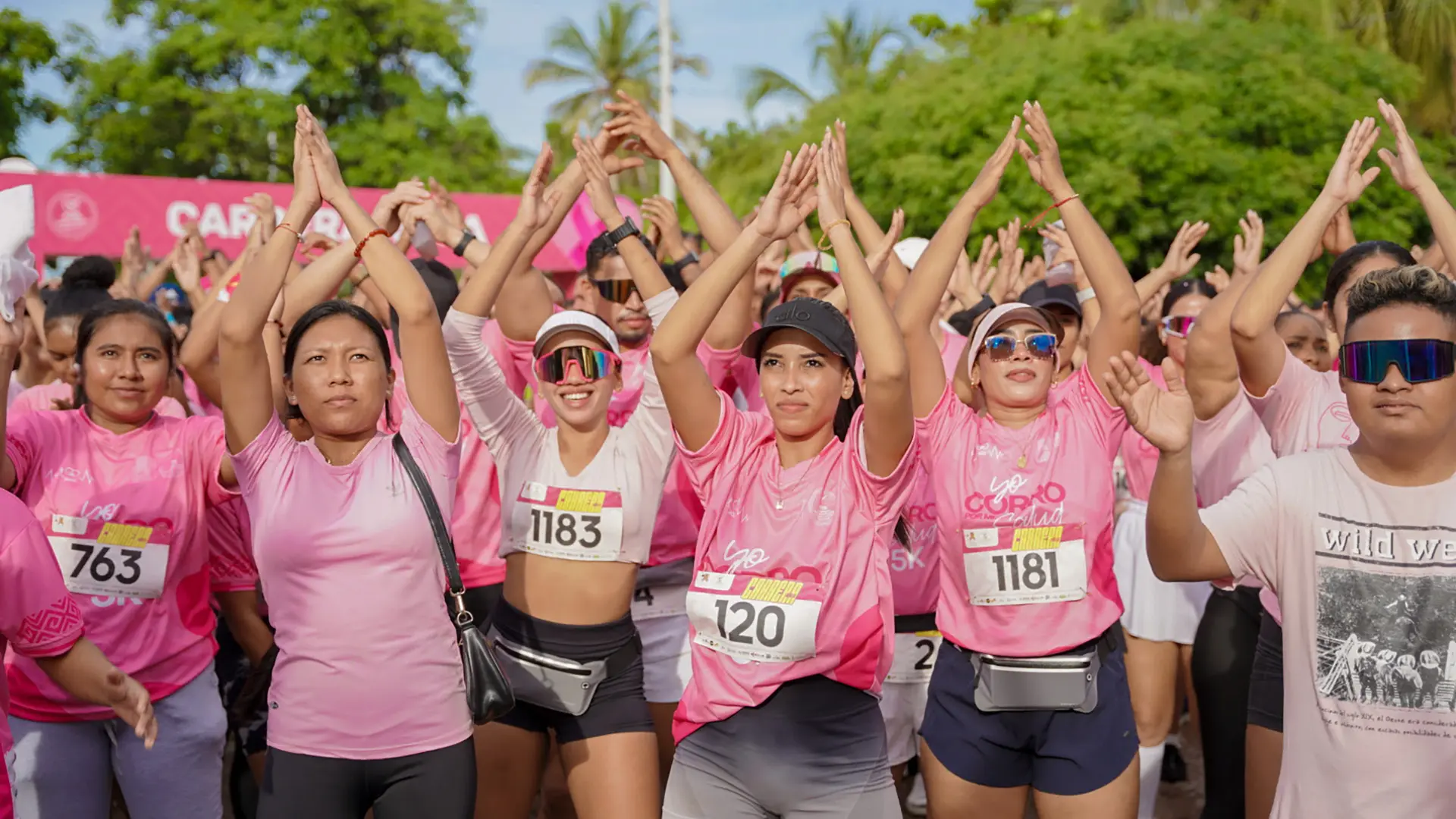 Grupo de participantes con camisetas rosas reunidos bajo un arco que dice “Carrera Rosa”, listos para iniciar una caminata o carrera en apoyo a la lucha contra el cáncer de mama.