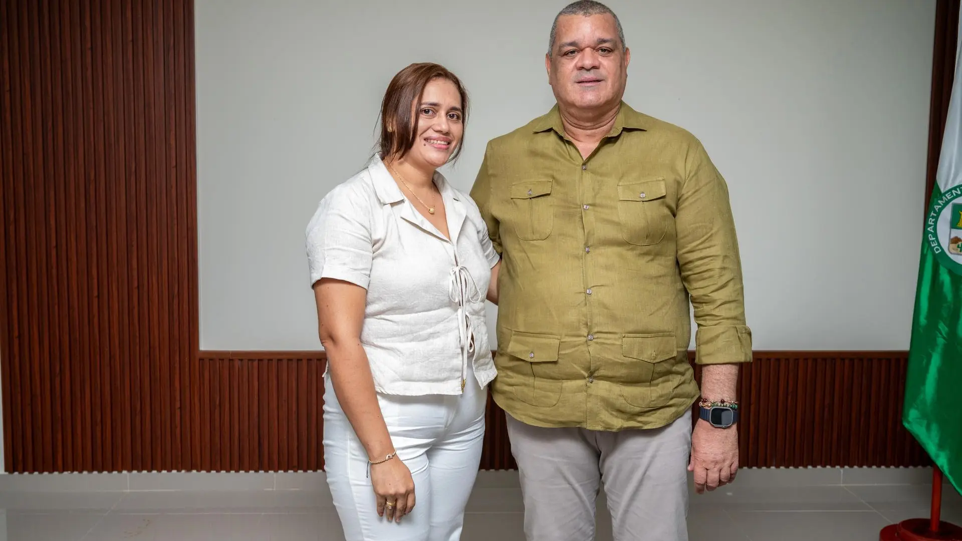 Una mujer y un hombre posan sonrientes en un espacio institucional con fondo de madera y una bandera verde del departamento de La Guajira . Ambos visten de manera formal y proyectan una imagen de colaboración y liderazgo académico