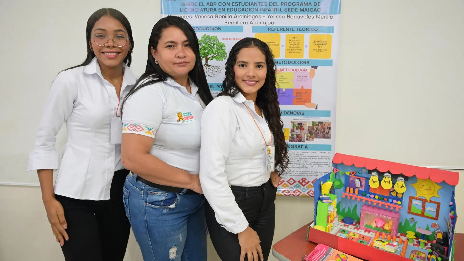 Tres mujeres posan sonrientes frente a un cartel académico que presenta un proyecto del programa de Licenciatura en Educación Infantil de la Universidad de La Guajira, Sede Maicao. Sobre una mesa se exhibe una maqueta colorida que representa un aula escolar. El cartel al fondo muestra el título del trabajo investigativo y el logotipo institucional.