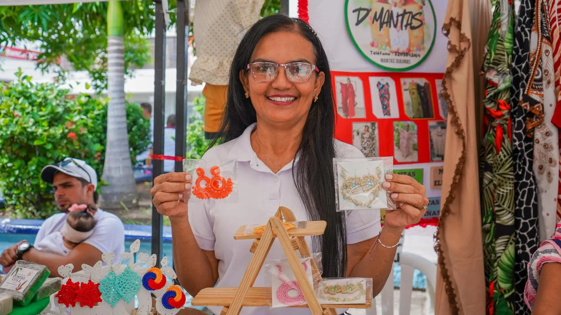 Mujer sonriente exhibe bisutería artesanal en un puesto de feria, sosteniendo un par de aretes tejidos mientras muestra otros accesorios en exposición.