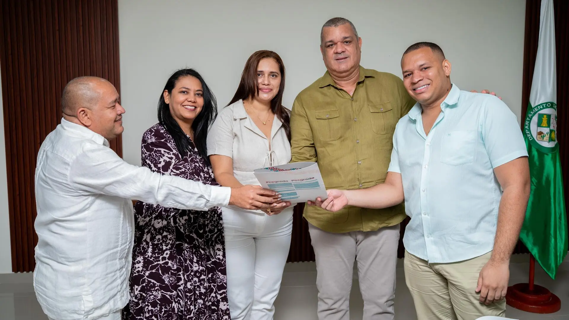 Cinco personas ,tres hombres y dos mujeres sostienen un documento con el logo de la Universidad de La Guajira. La escena refleja trabajo en equipo y compromiso institucional en un ambiente académico formal