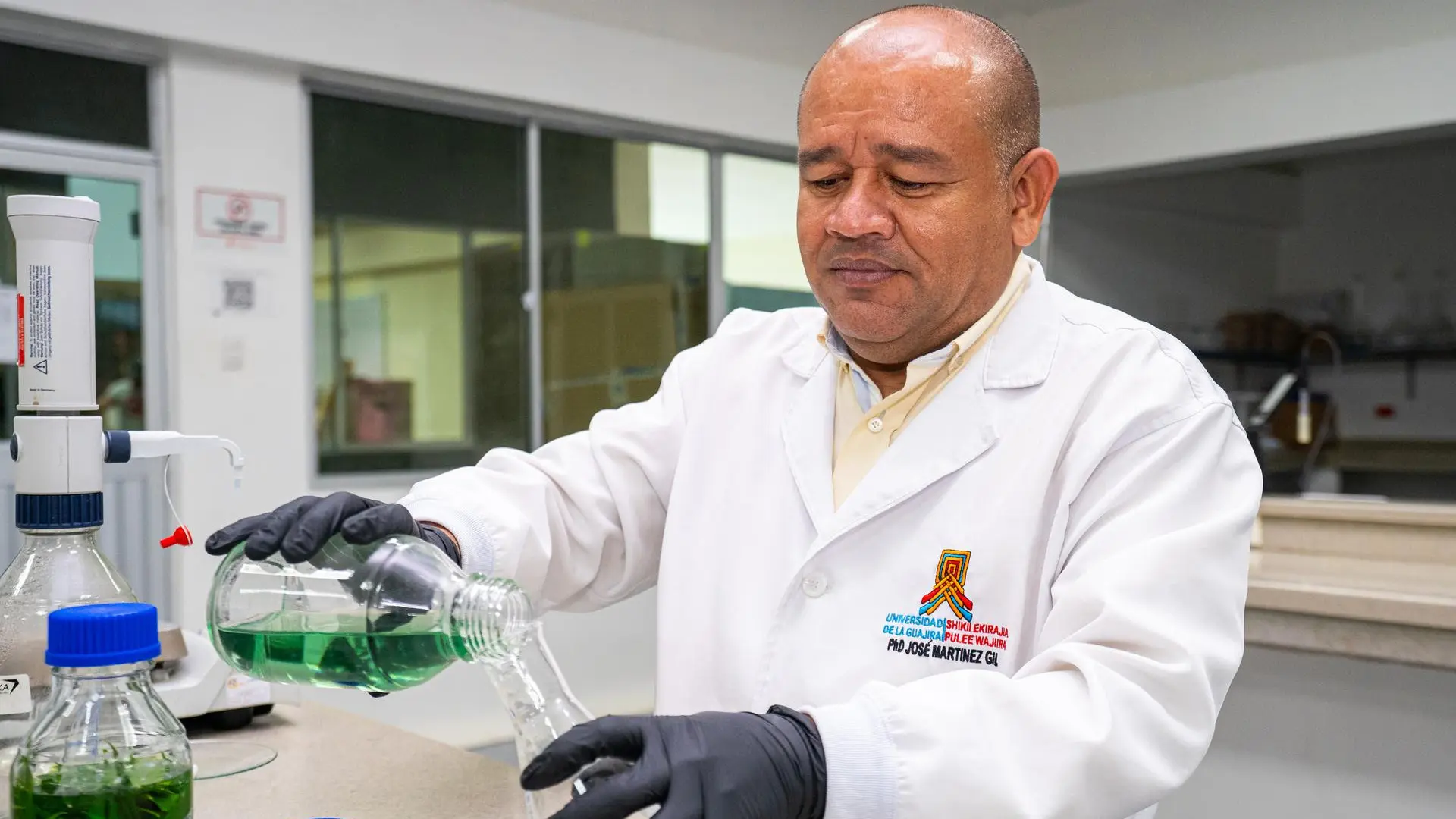 José Manuel Martinez, docente investigador de Uniguajira en un laboratorio. Viste una bata blanca con el logo institucional y su nombre bordado, y usa guantes negros mientras vierte un líquido verde de un frasco a otro sobre una mesa de trabajo. En el entorno se observan equipos y recipientes de laboratorio.