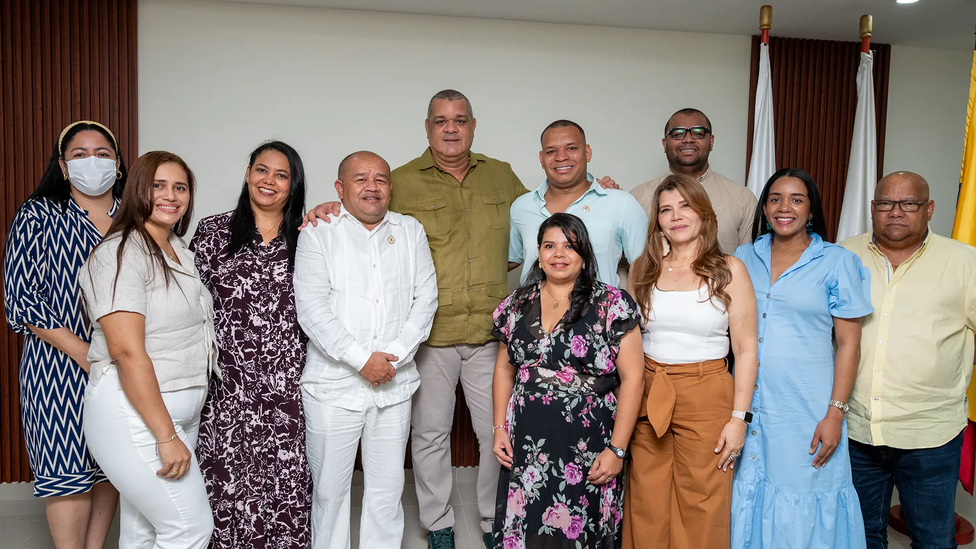Grupo de once personas adultas posando para una fotografía en un salón institucional. Están organizadas en dos filas: una fila frontal con mujeres y hombres de pie, y detrás otra fila con cuatro personas. Visten ropa formal y semiformal en colores variados. Al fondo se observan banderas y un muro con paneles de madera verticales